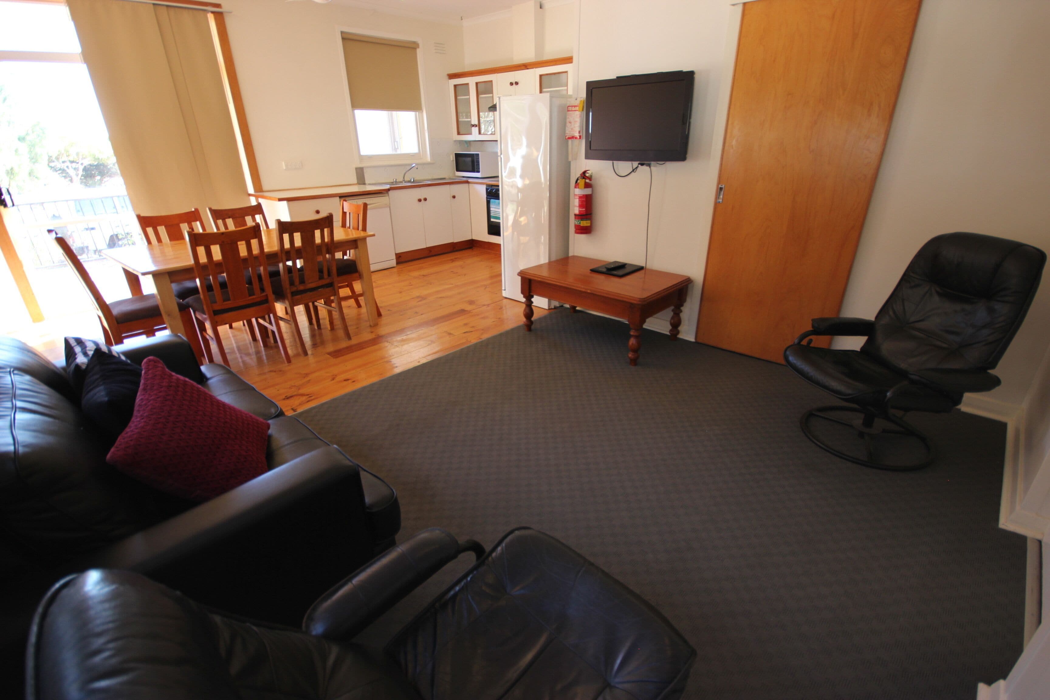 A living room with a black leather sofa, a dining table with chairs, a kitchen area, and a black leather chair.