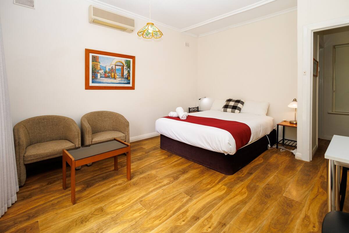 Une chambre à coucher avec un lit, deux fauteuils, un plancher en bois, un tableau au mur et un luminaire suspendu.