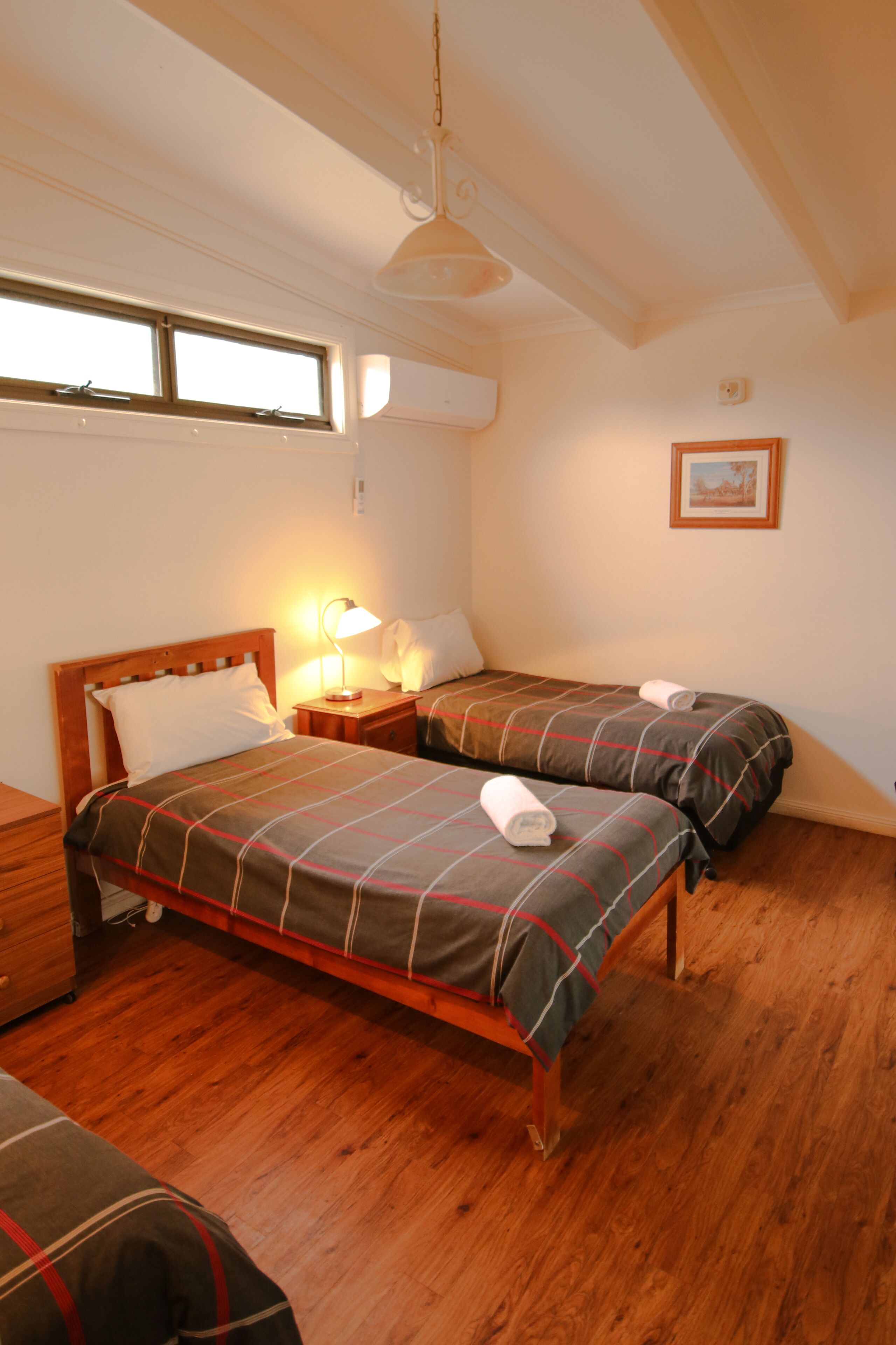 Un dormitorio con dos camas, piso de madera, ventilador de techo, una ventana y un cuadro en la pared.