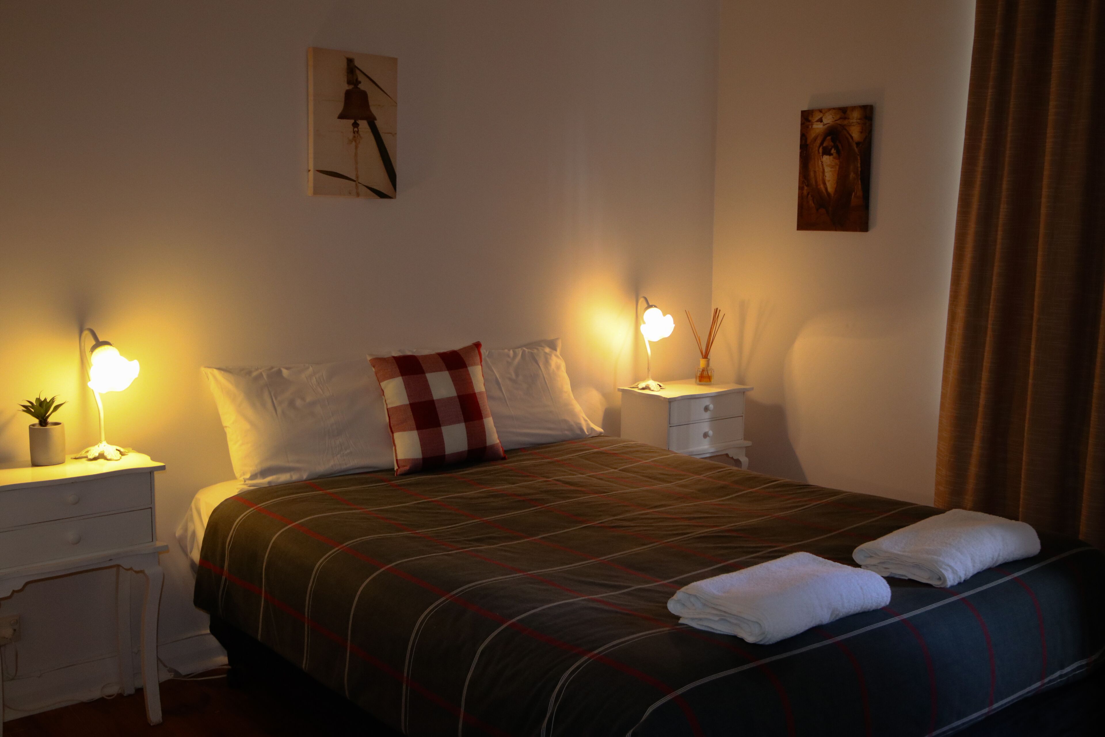 Una cama bien hecha con una colcha escocesa, dos almohadas blancas y dos toallas dobladas en el reposapiés. Hay mesitas de noche con lámparas y una pequeña planta en maceta a un lado.