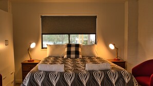 Un lit bien fait, recouvert d’une couvre-lit à motifs, deux lampes de chevet, une fenêtre donnant sur un beau paysage et un fauteuil rouge.