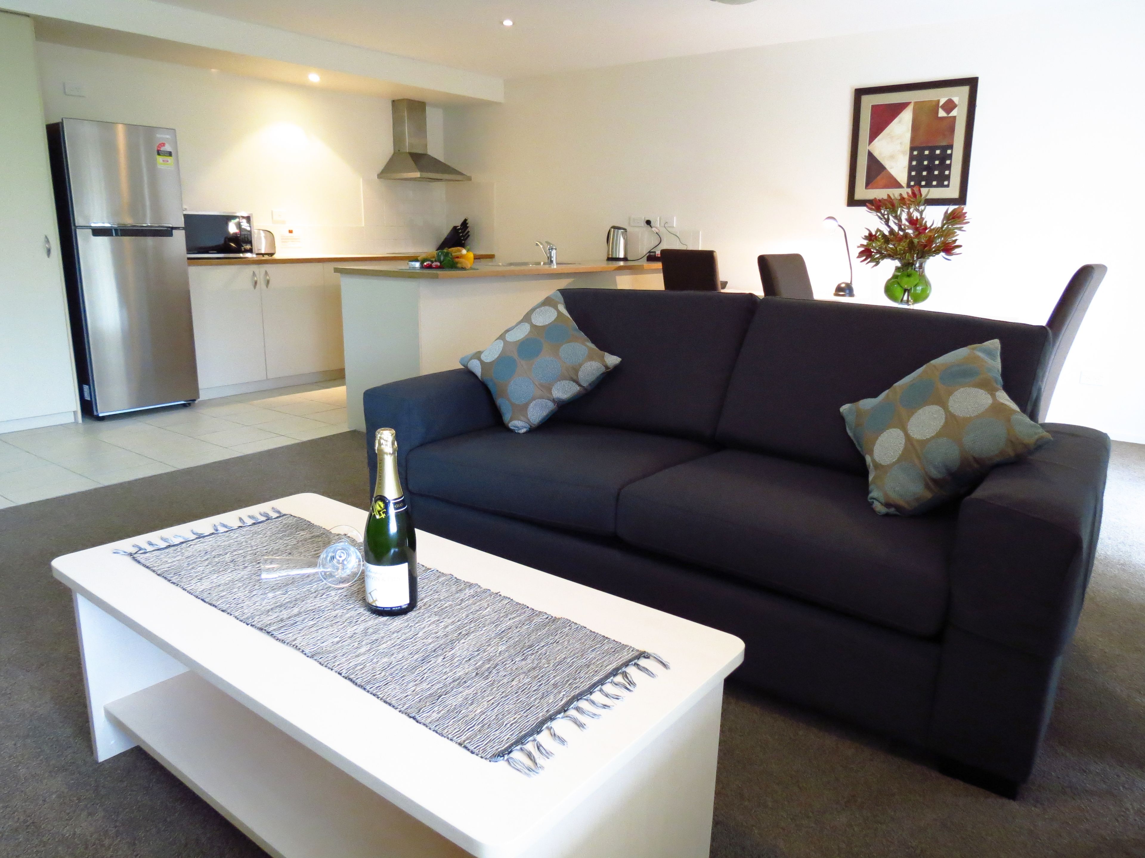 A modern living room with a dark sofa, a white coffee table with a bottle, and a kitchen in the background.