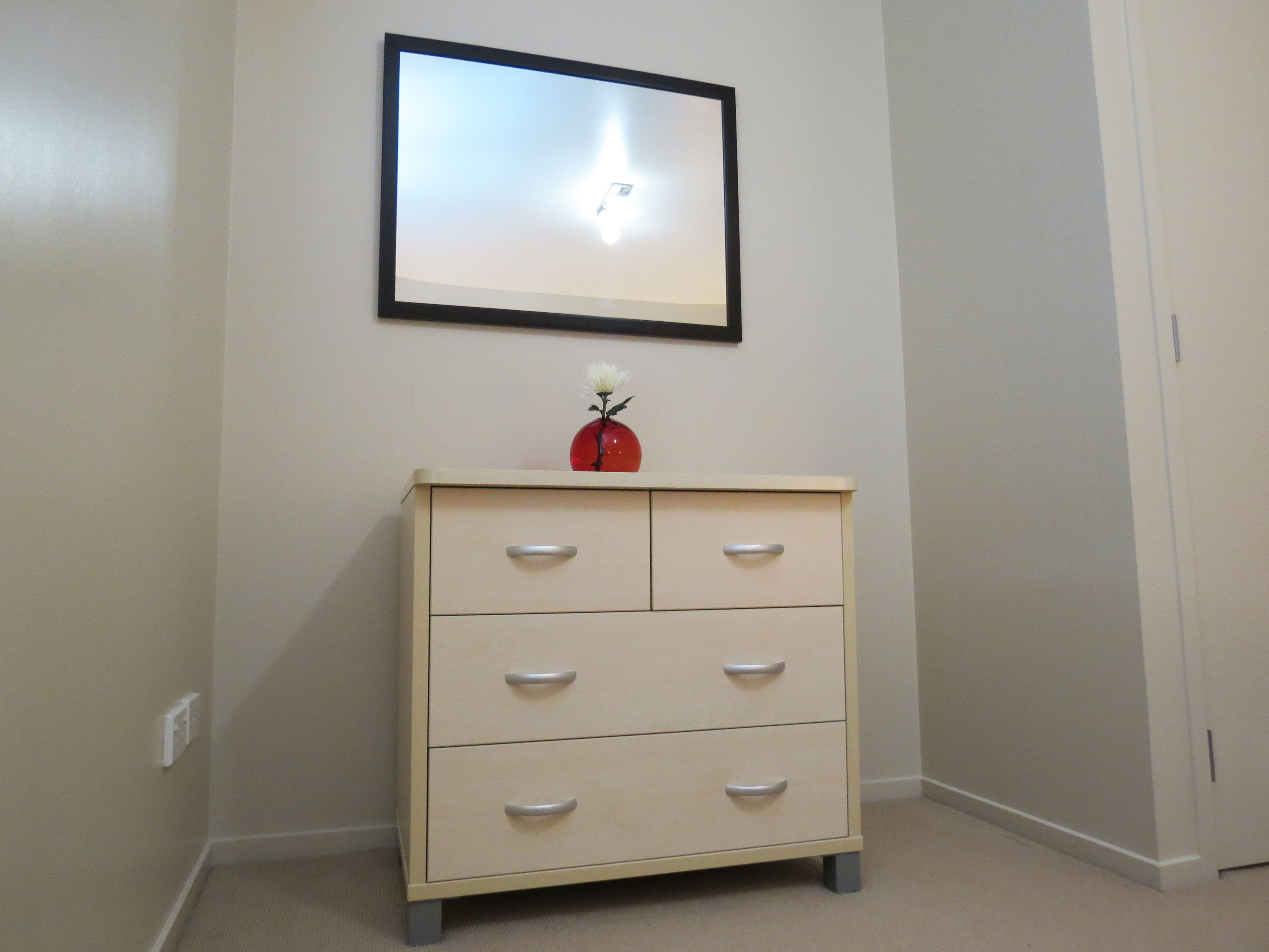 A small, cream-colored dresser with four drawers and a single vase with a white flower on top, positioned against a plain wall with a framed picture above it.