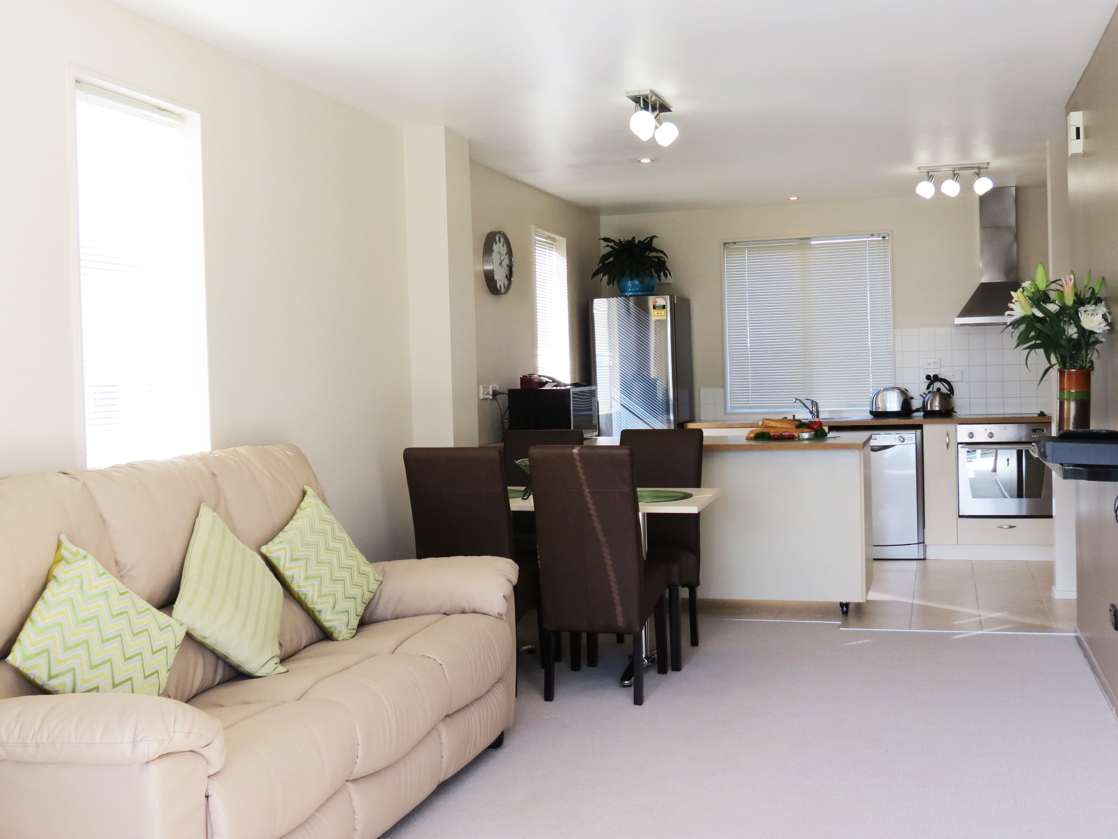 A modern living room with a beige sofa, a dining table with chairs, a kitchen area with appliances, and a large window with blinds.
