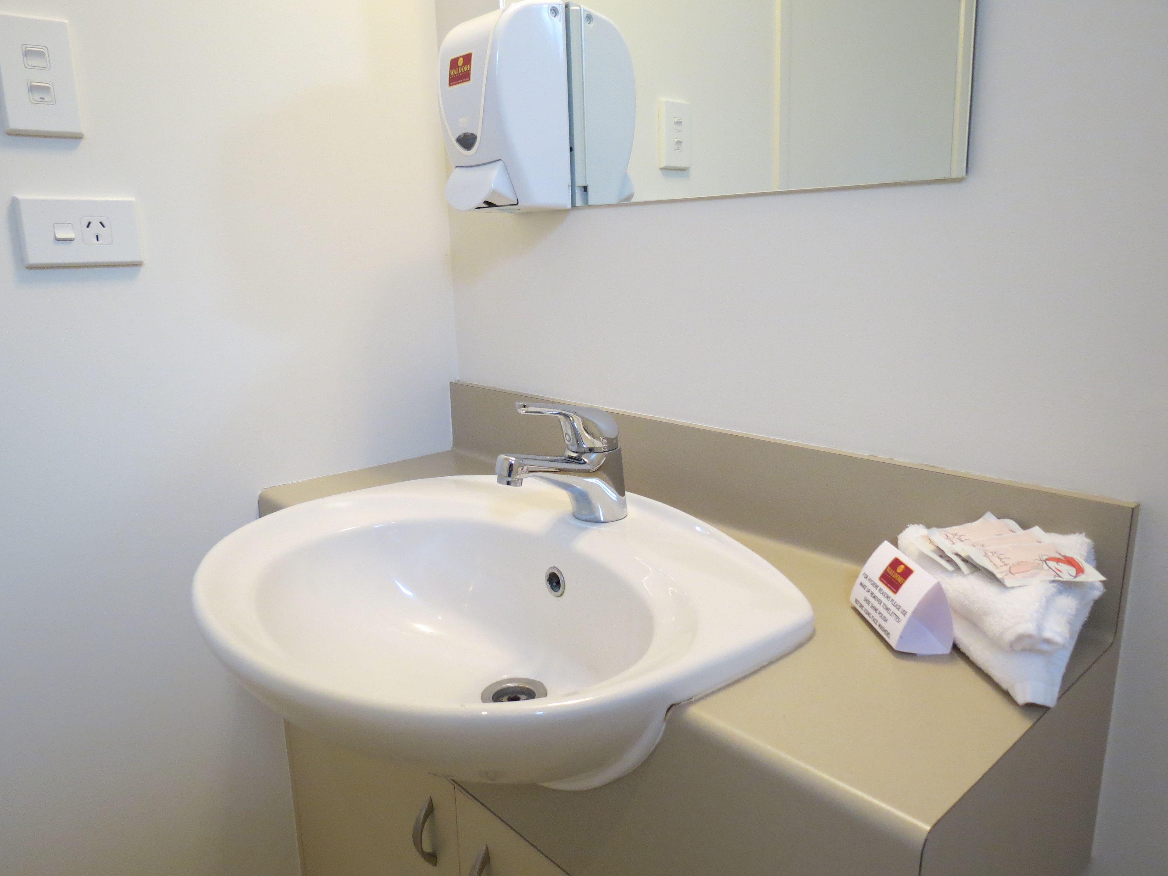 A white bathroom sink with a mirror above it, a towel dispenser, and a beige countertop.