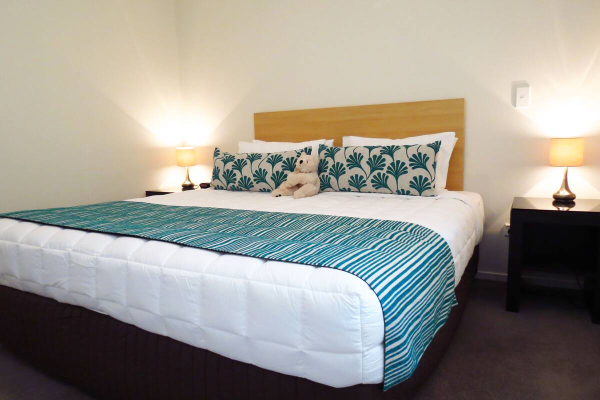 Una cama impecablemente arreglada con colcha blanca y azul estampada, dos almohadas a juego y un cabecero de madera. Hay dos mesitas de noche con lámparas, una a cada lado de la cama.