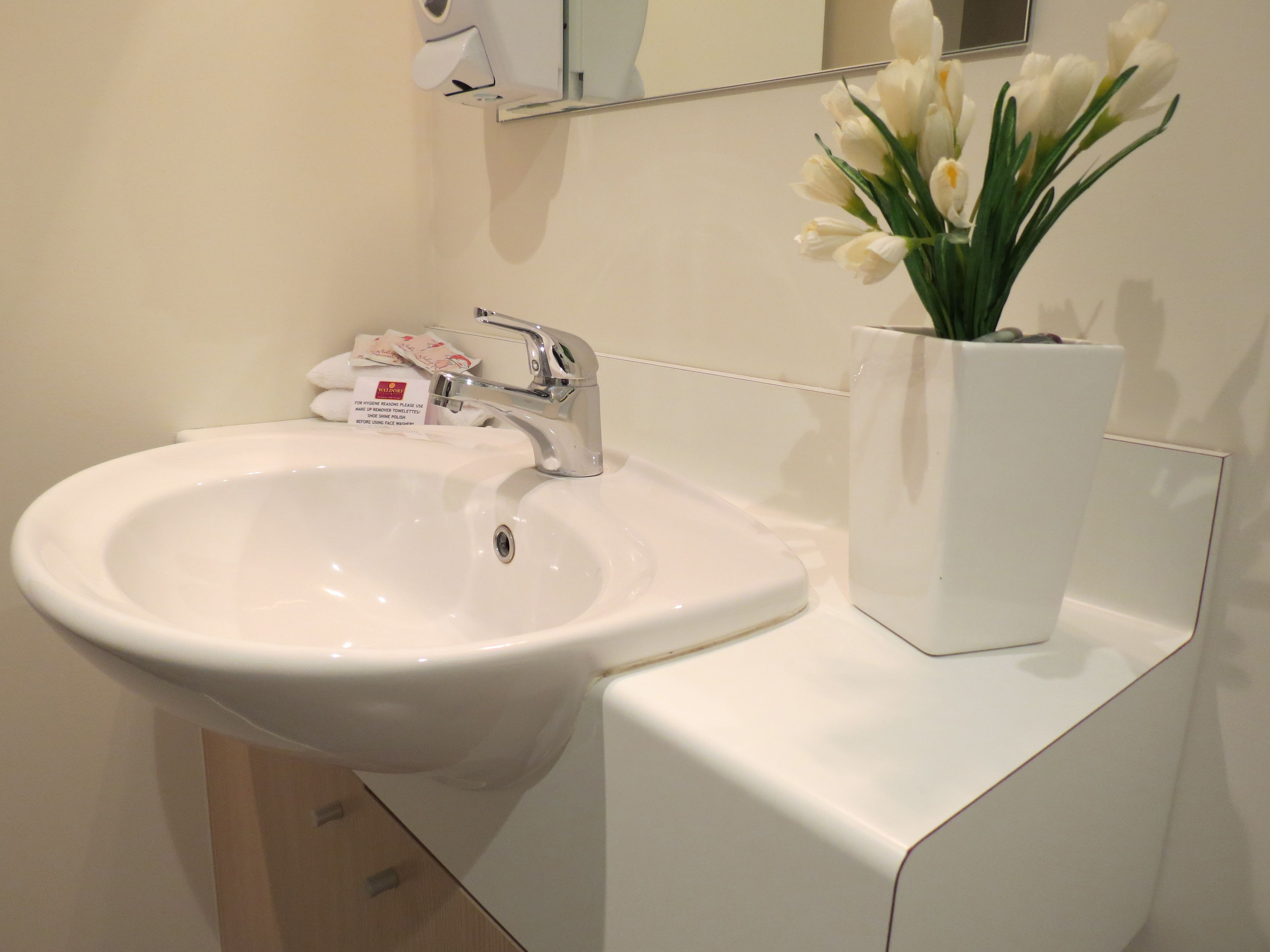 A white bathroom sink with a chrome faucet, a white vase with white flowers, and a hand soap dispenser on the counter.