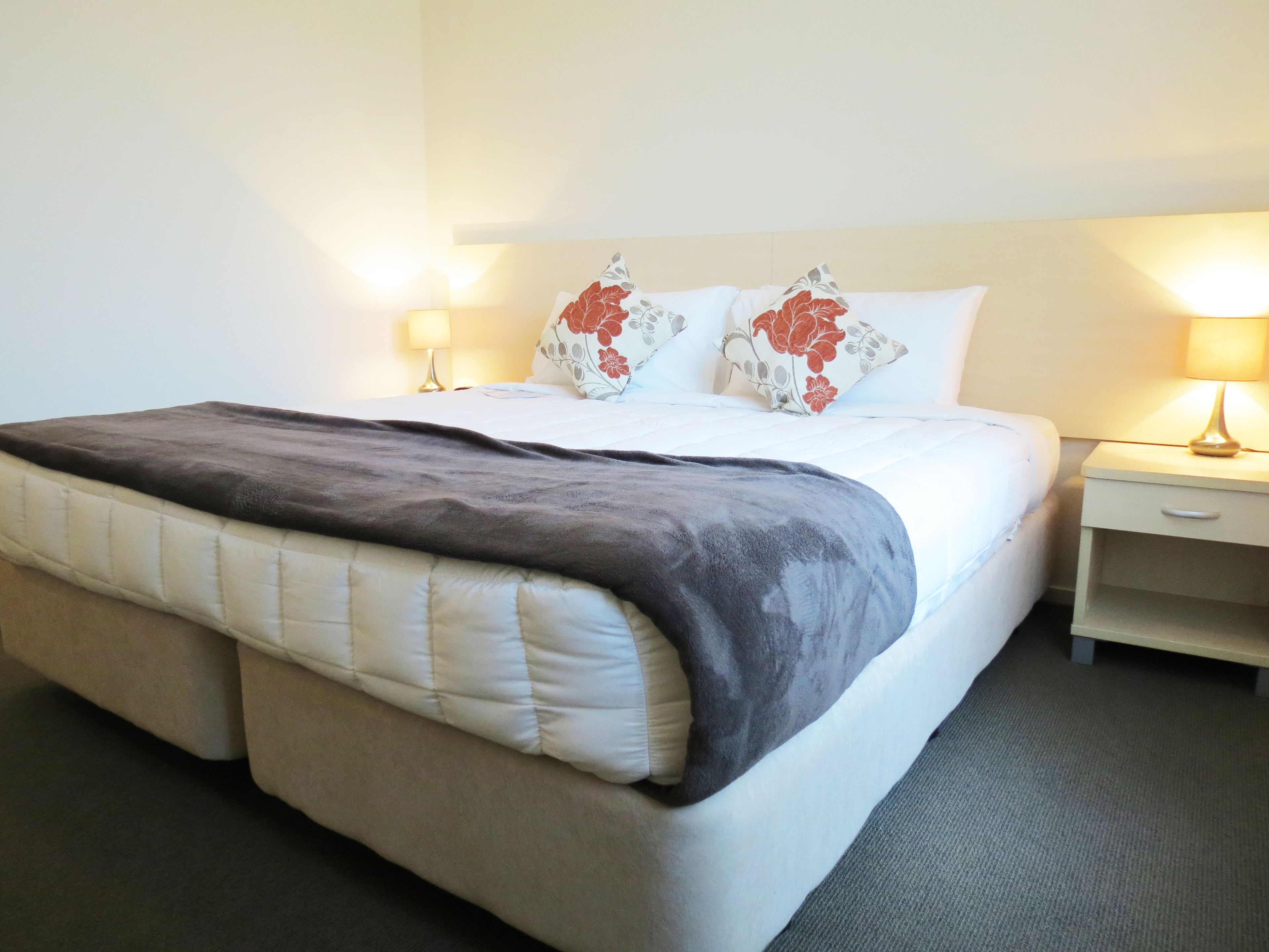 A neatly made bed with a grey blanket, white pillows with floral patterns, and two bedside lamps on either side.