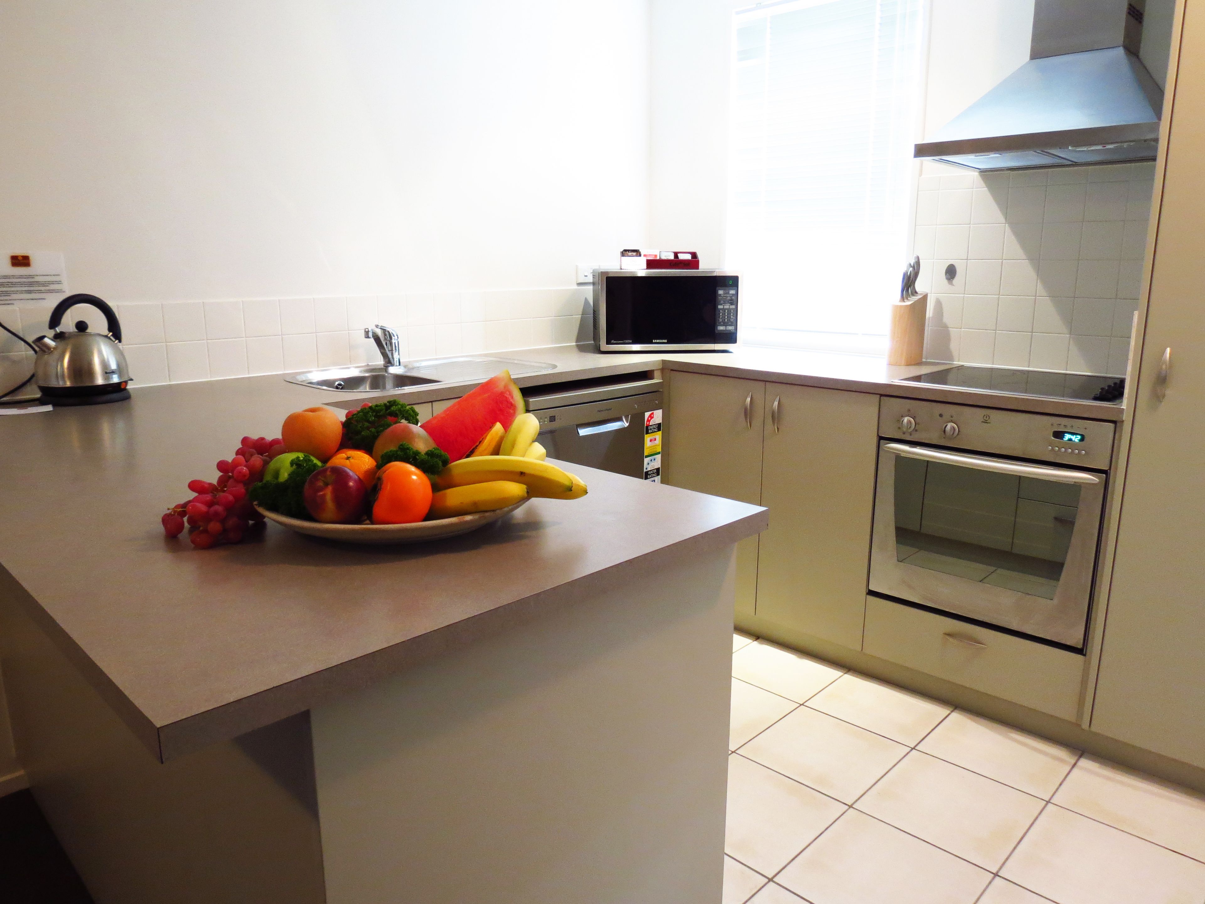 A kitchen with a fruit bowl on the counter, a kettle, a microwave, and an oven.