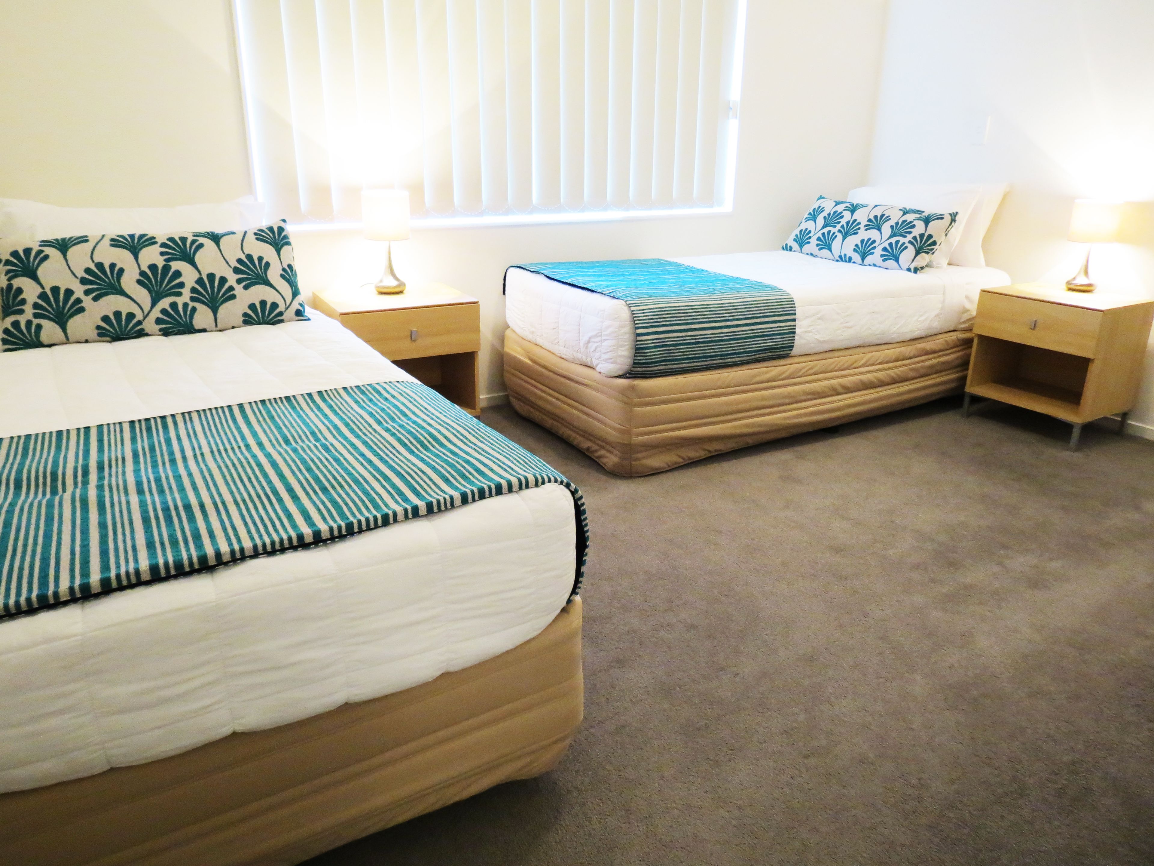 A hotel room with two beds, each with a different patterned bedspread. There are bedside tables with lamps and a window with blinds.