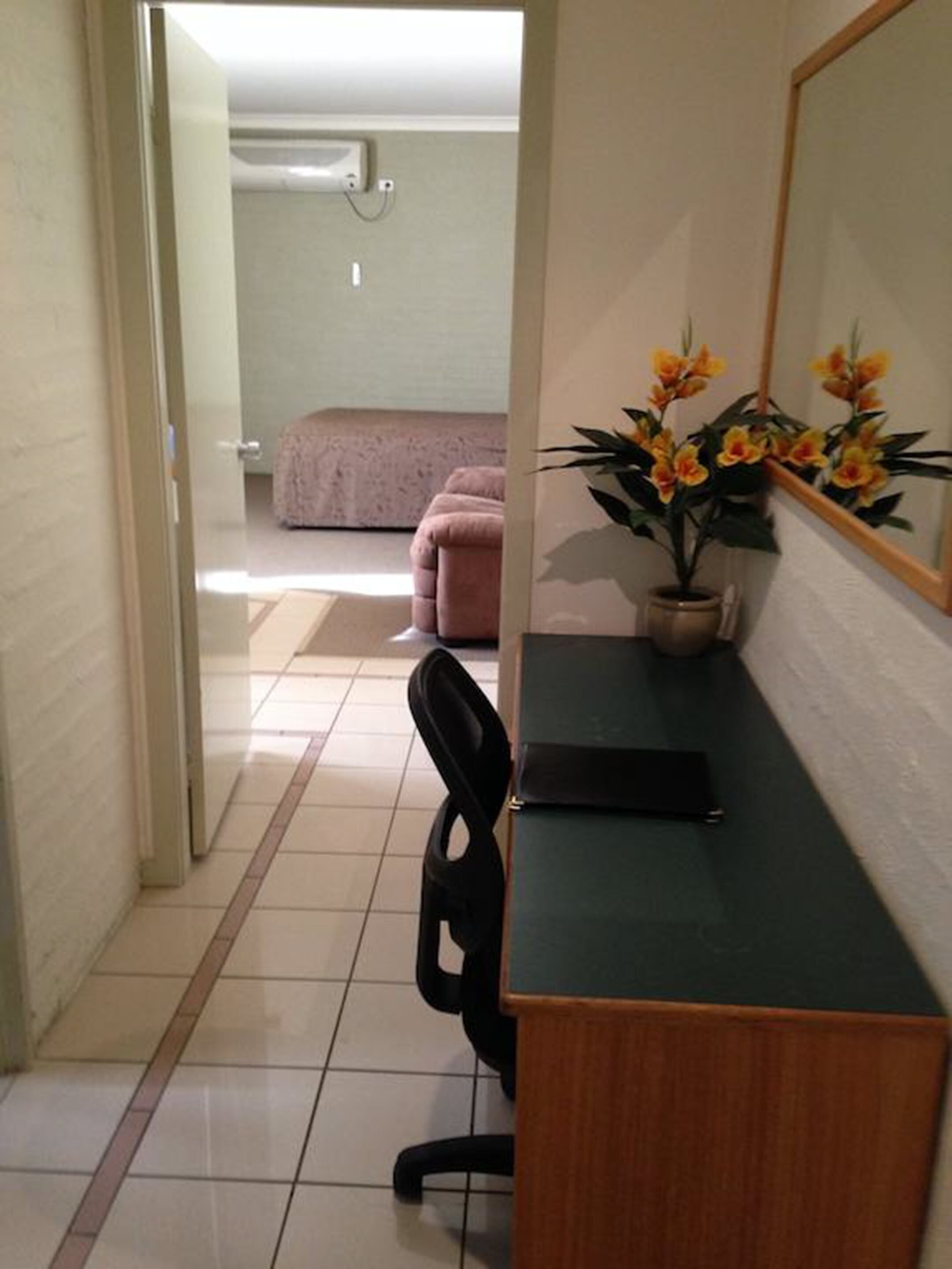 A hotel room with a desk, chair, potted plant, and a bed in the background.