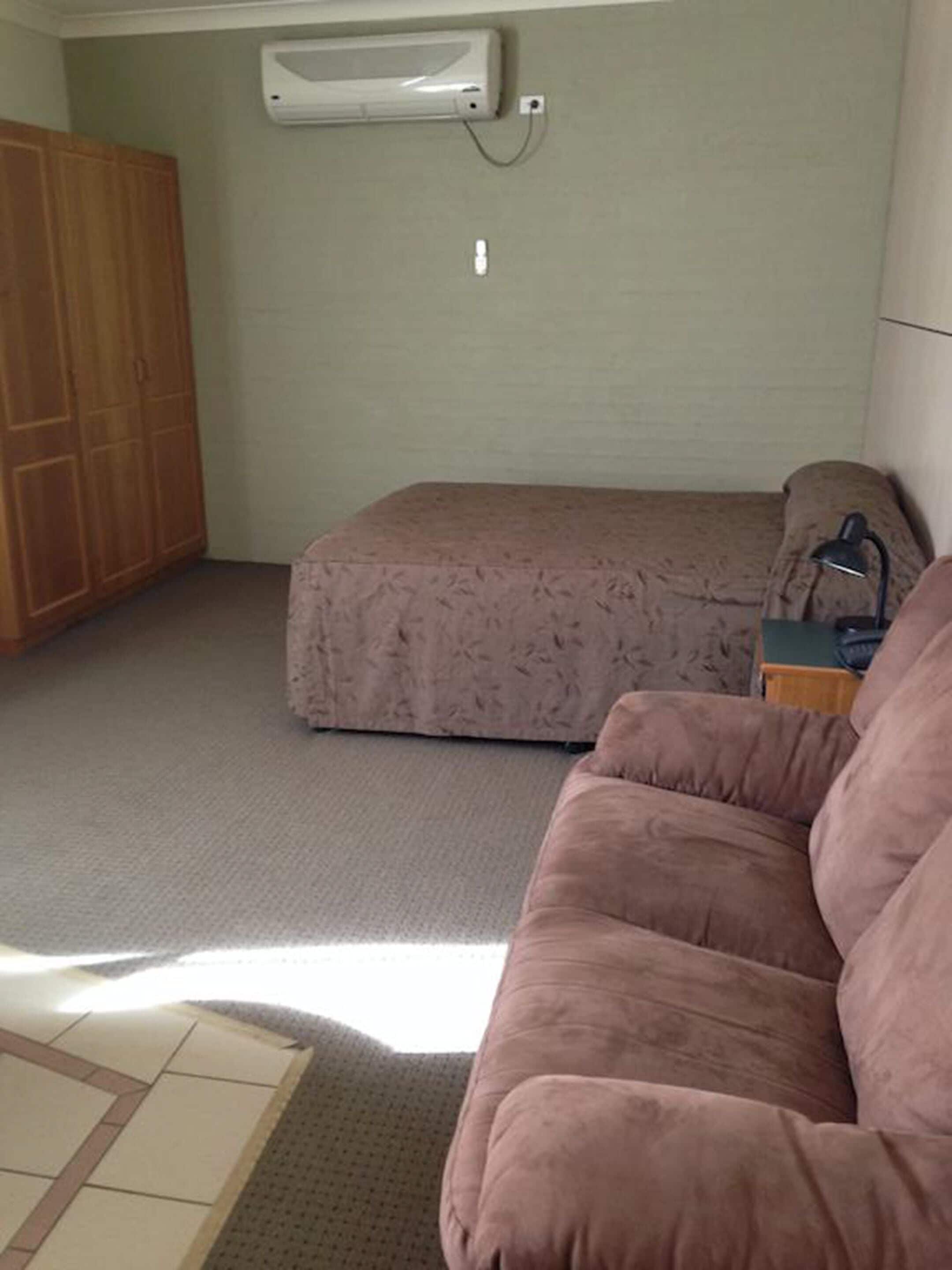 A room with a bed, a sofa, a wardrobe, and an air conditioning unit on the wall.