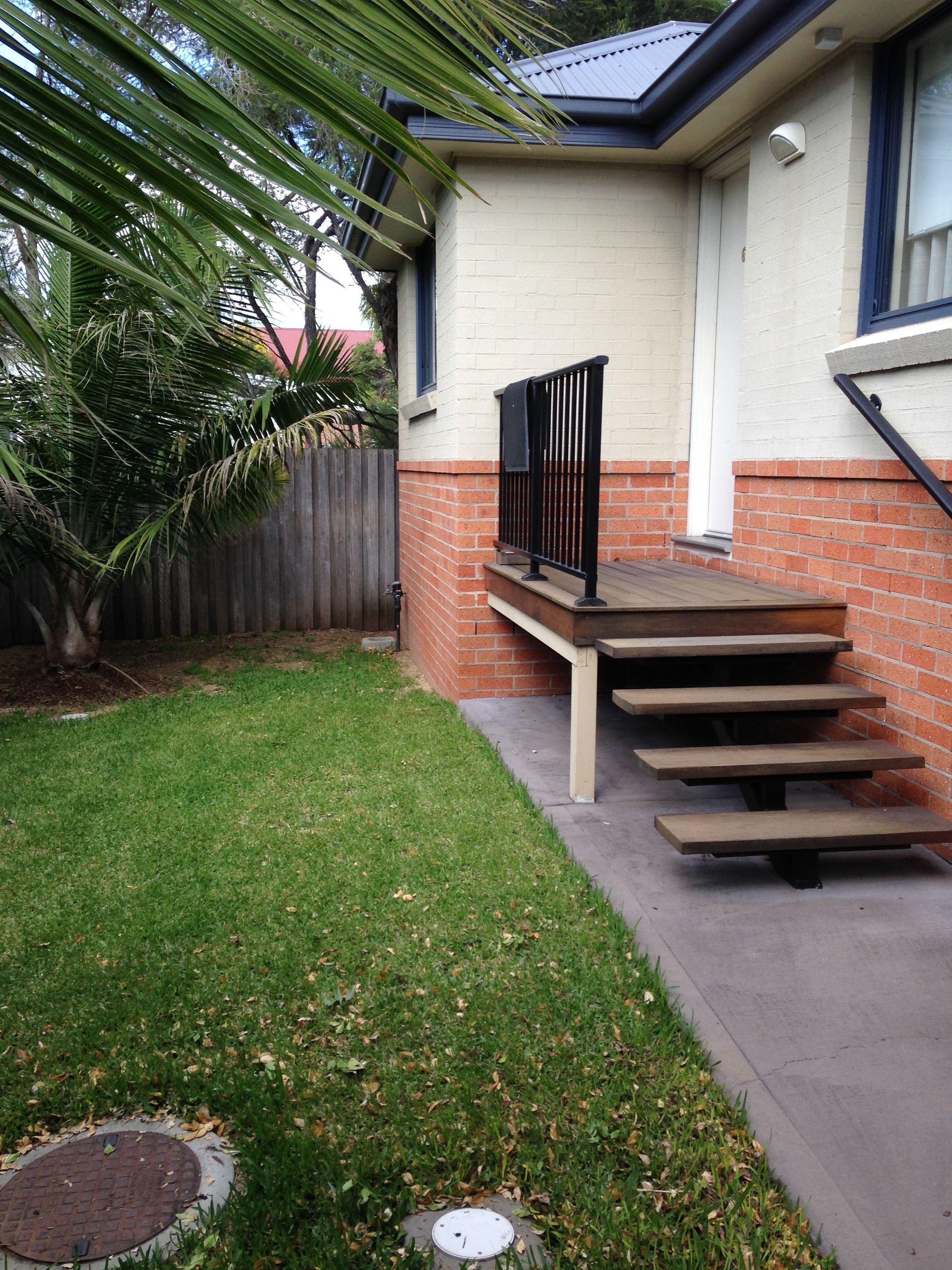 Una propiedad residencial con una escalera de hormigón que conduce a una terraza de madera, rodeada de césped cuidado y una valla de madera.