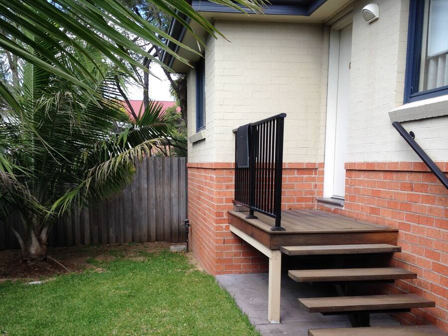 Een woonhuis met een betonnen trap die leidt naar een houten terras, omgeven door een verzorgd gazon en een houten omheining.