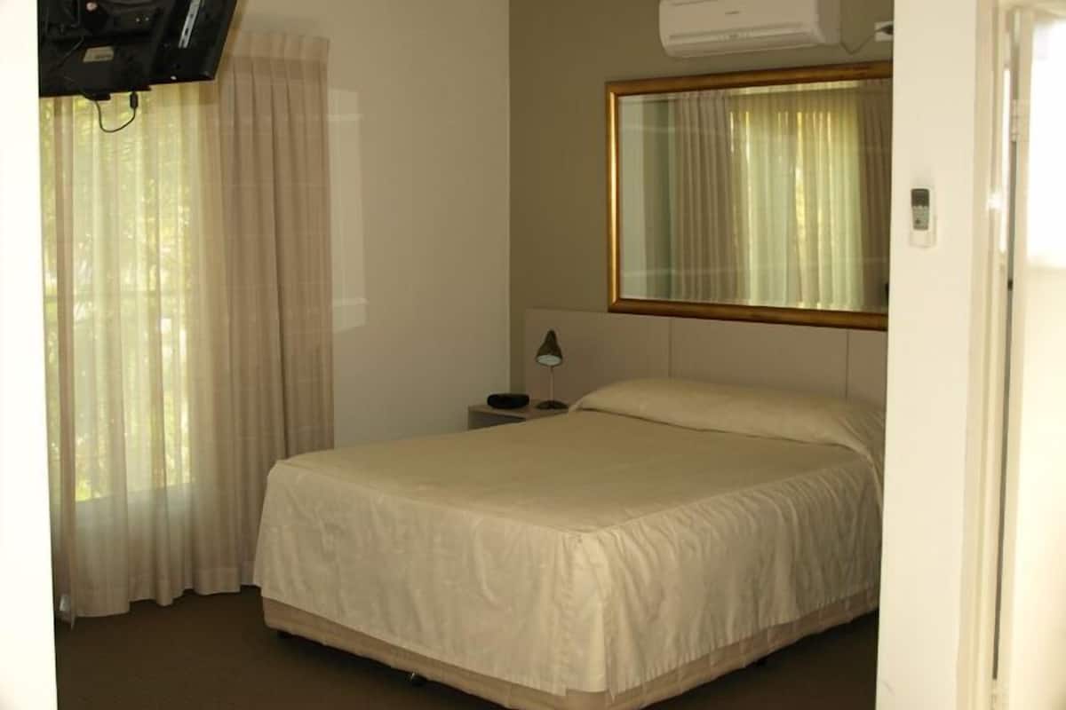Una habitación de hotel con una cama, una mesita de noche, una lámpara, una ventana con cortinas y un equipo de aire acondicionado.