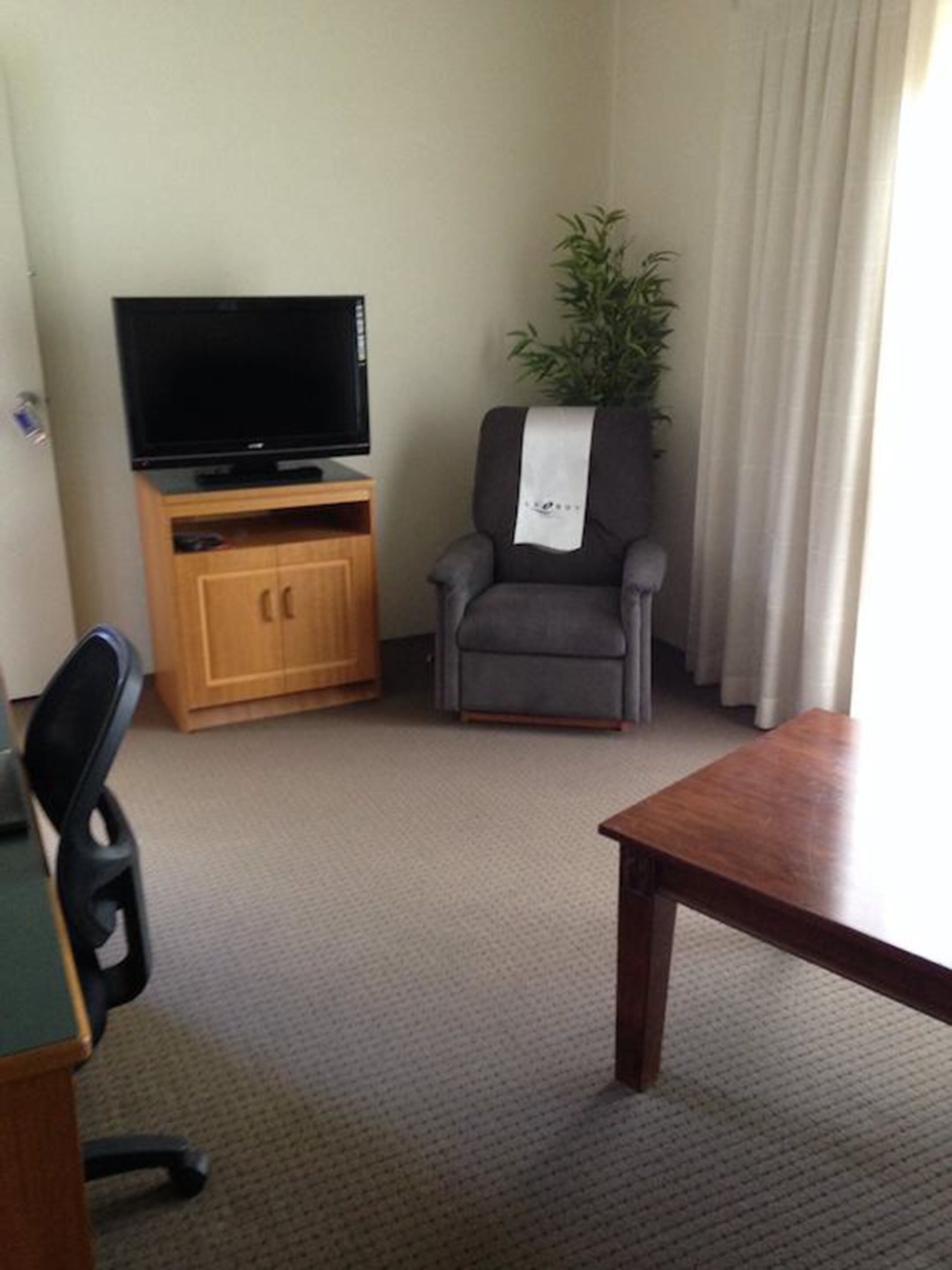 A hotel room with a wooden entertainment unit, a television, a grey armchair with a white towel, a small table, and a potted plant.