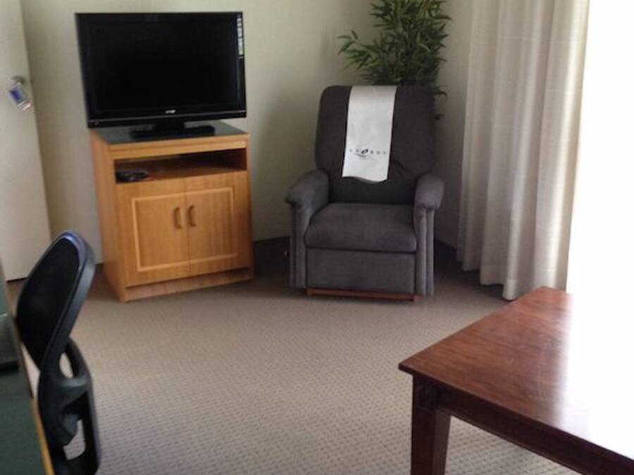 A hotel room with a wooden entertainment unit, a television, a grey armchair with a white towel, a small table, and a potted plant.