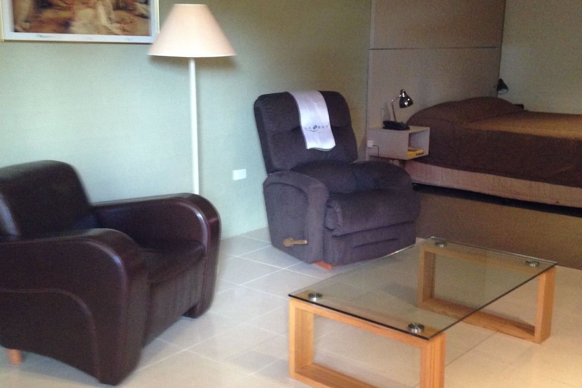 Una sala de estar con sofá, sillón reclinable, mesa de centro de vidrio, lámpara y cama.