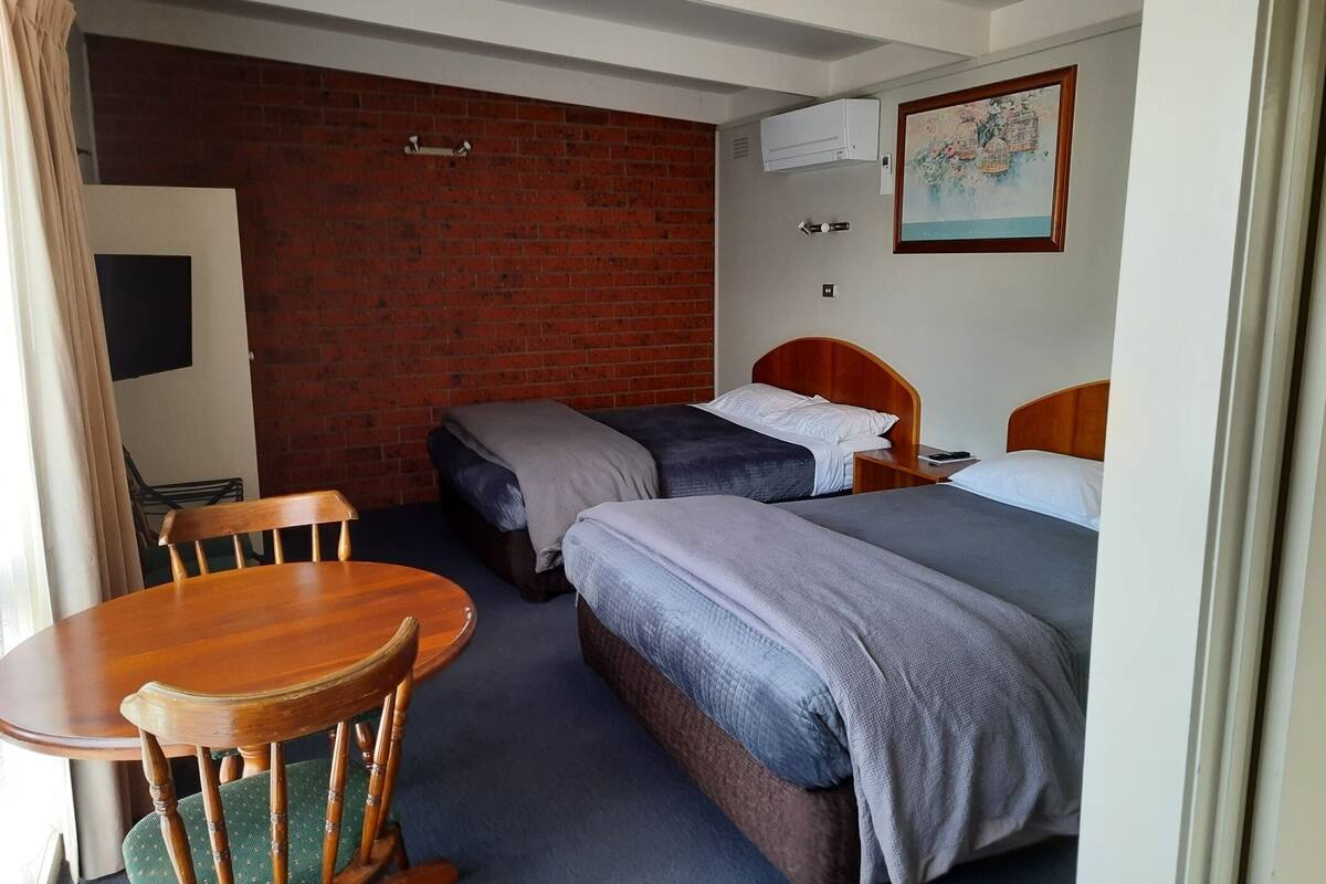 Habitación de hotel con dos camas individuales, una mesa de madera con una silla, un televisor y una pared de ladrillo.
