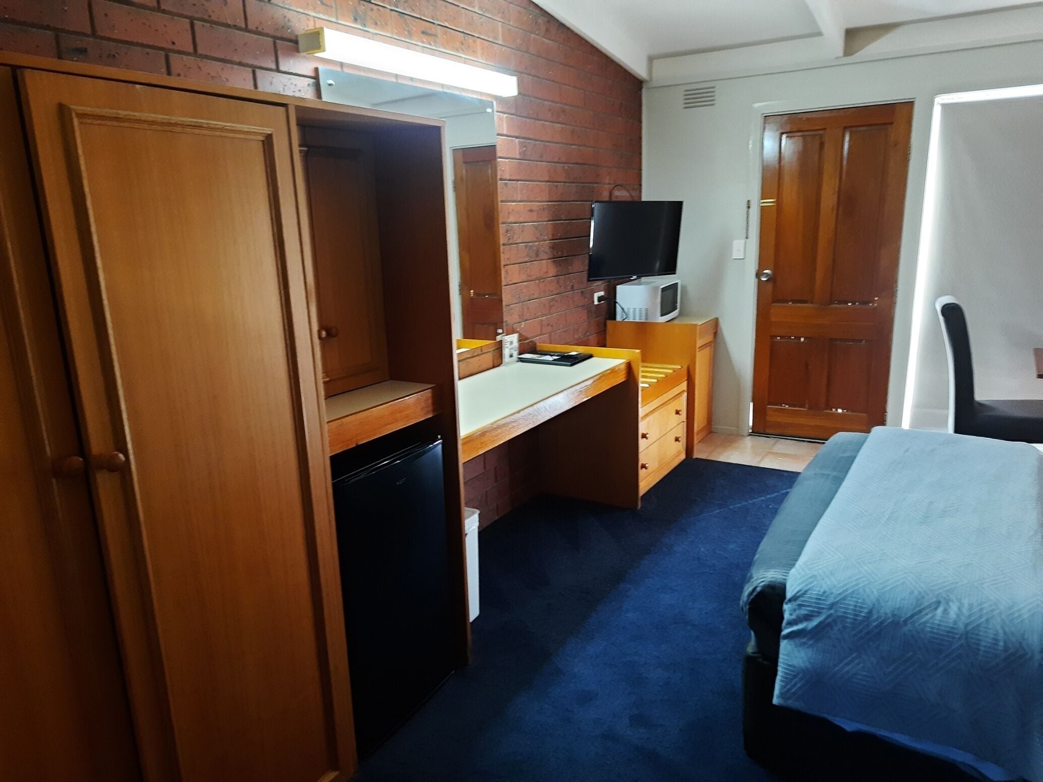 A compact hotel room with a wooden wardrobe, a desk with a microwave, a television, and a bed with a blue cover.