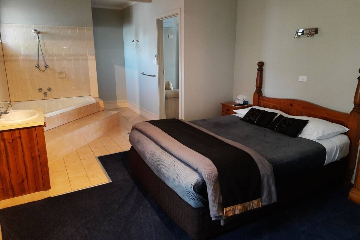 Un dormitorio con una cama grande, cabecera de madera, una mesita de noche y un baño con bañera y ducha.