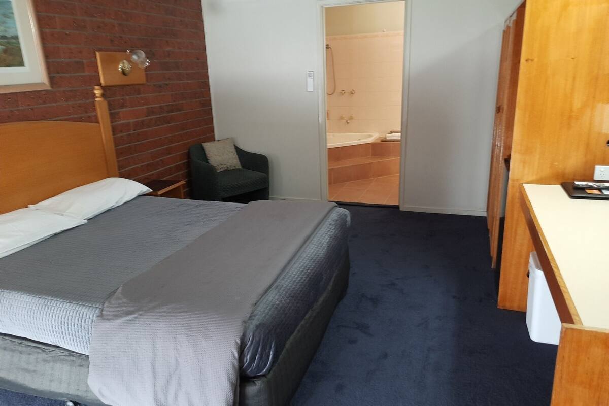 Habitación de hotel con cama, una silla, un escritorio y un baño visible a través de una puerta abierta.