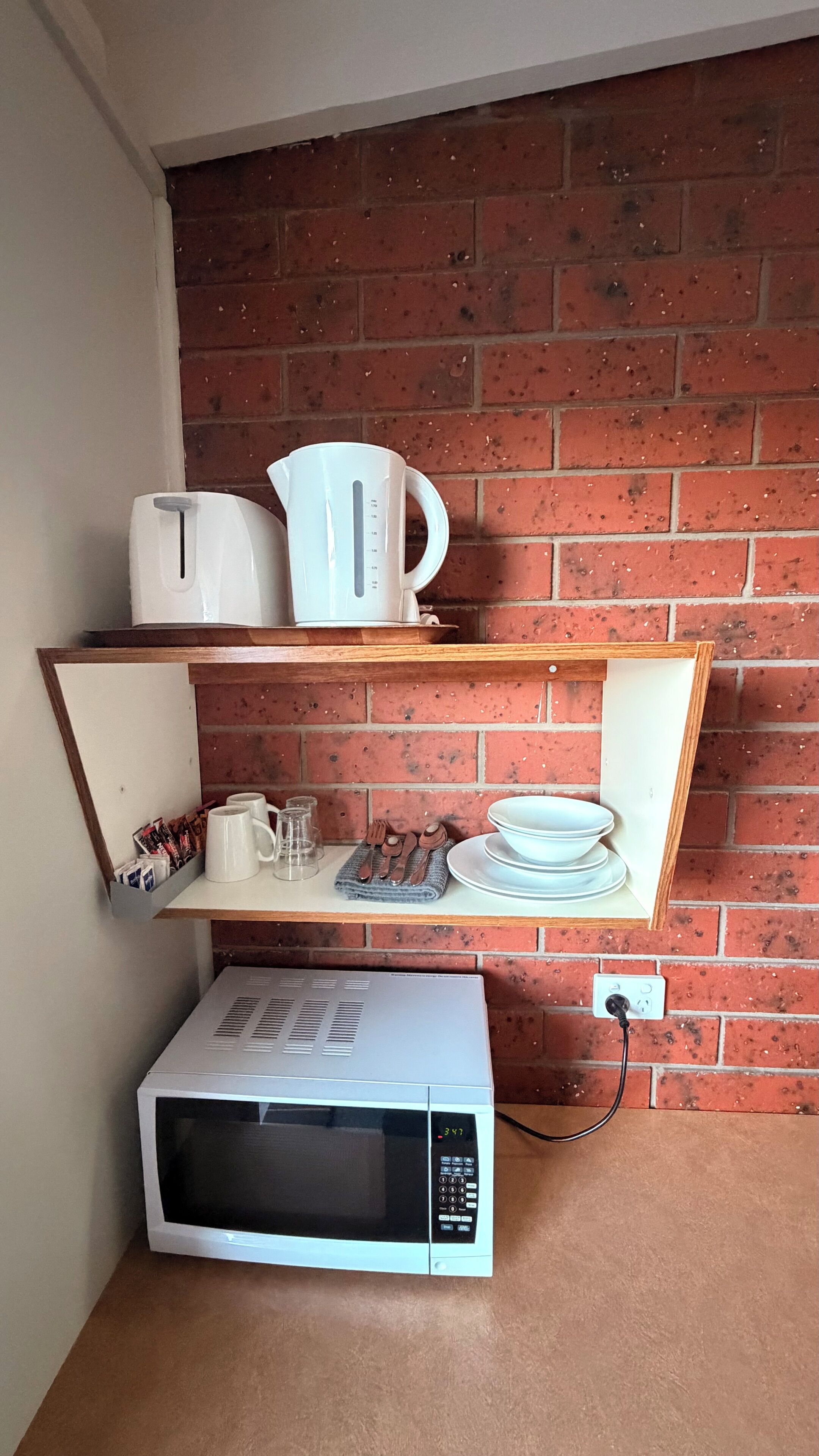 Una cocina con pared de ladrillo, microondas, hervidor y estante con tazas y platos.
