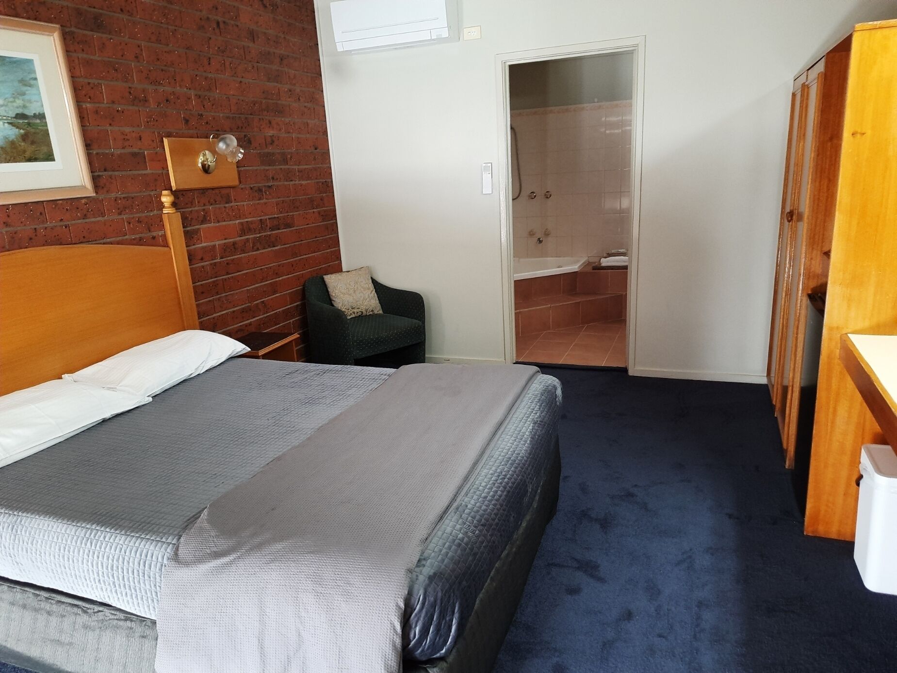A hotel room with a large bed, a chair, a wooden wardrobe, and a bathroom with a bathtub and shower.