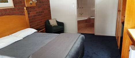 A hotel room with a large bed, a chair, a wooden wardrobe, and a bathroom with a bathtub and shower.