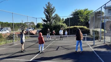 Ein Tennisplatz mit Netz, umgeben von einem Maschendrahtzaun, Bäumen und geparkten Autos im Hintergrund.