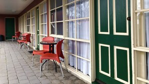 Uma área de estar coberta ao ar livre com cadeiras vermelhas e uma mesa, janelas amplas com molduras brancas e uma porta verde.