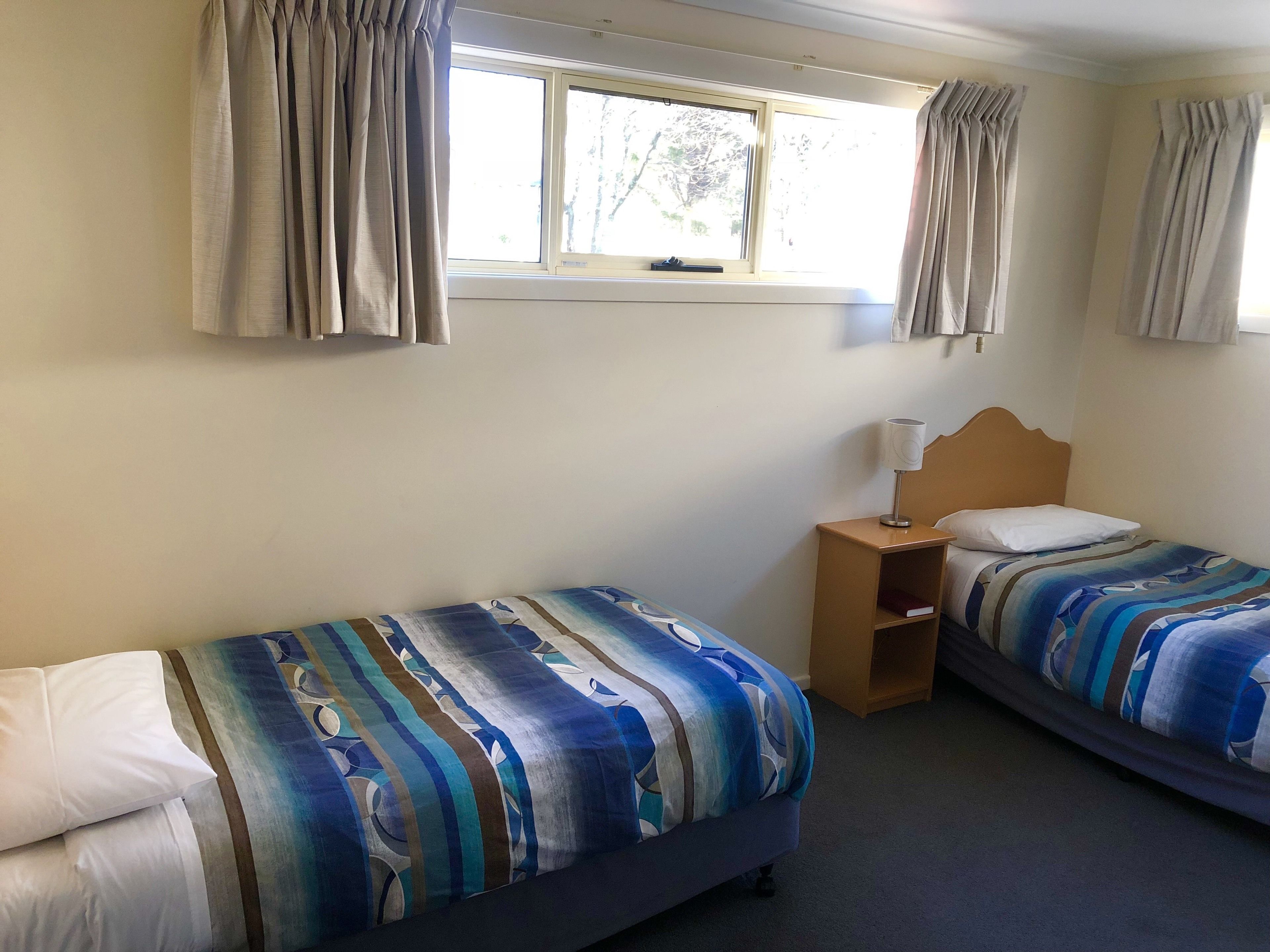 A room with two beds, a nightstand, a lamp, and a window with curtains.
