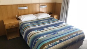 Quarto de hotel com cabeceira de madeira, cama com colcha listrada, mesa de cabeceira e criado-mudo.