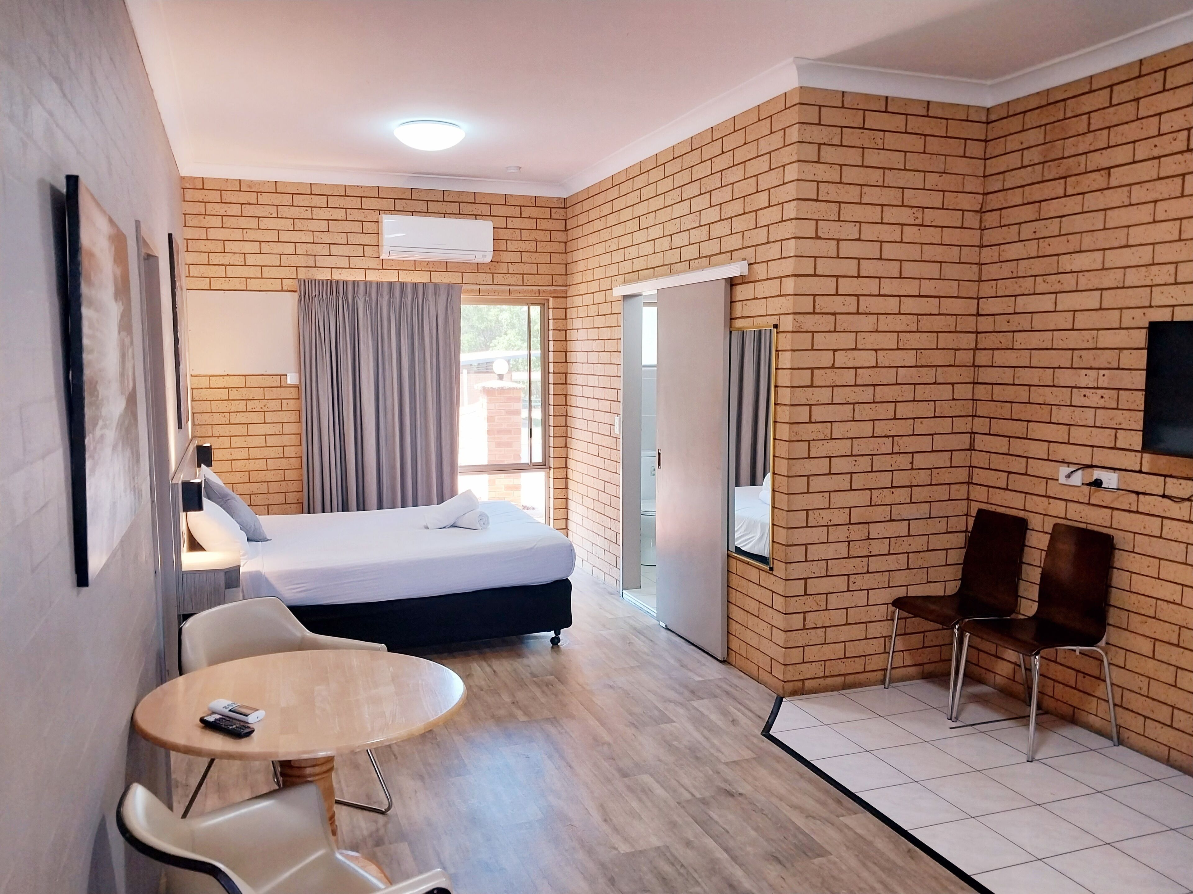 A hotel room with a brick wall, a bed, a desk with a chair, a small round table with a chair, and a TV mounted on the wall.