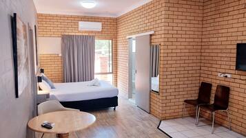 A hotel room with a brick wall, a bed, a desk with a chair, a small round table with a chair, and a TV mounted on the wall.