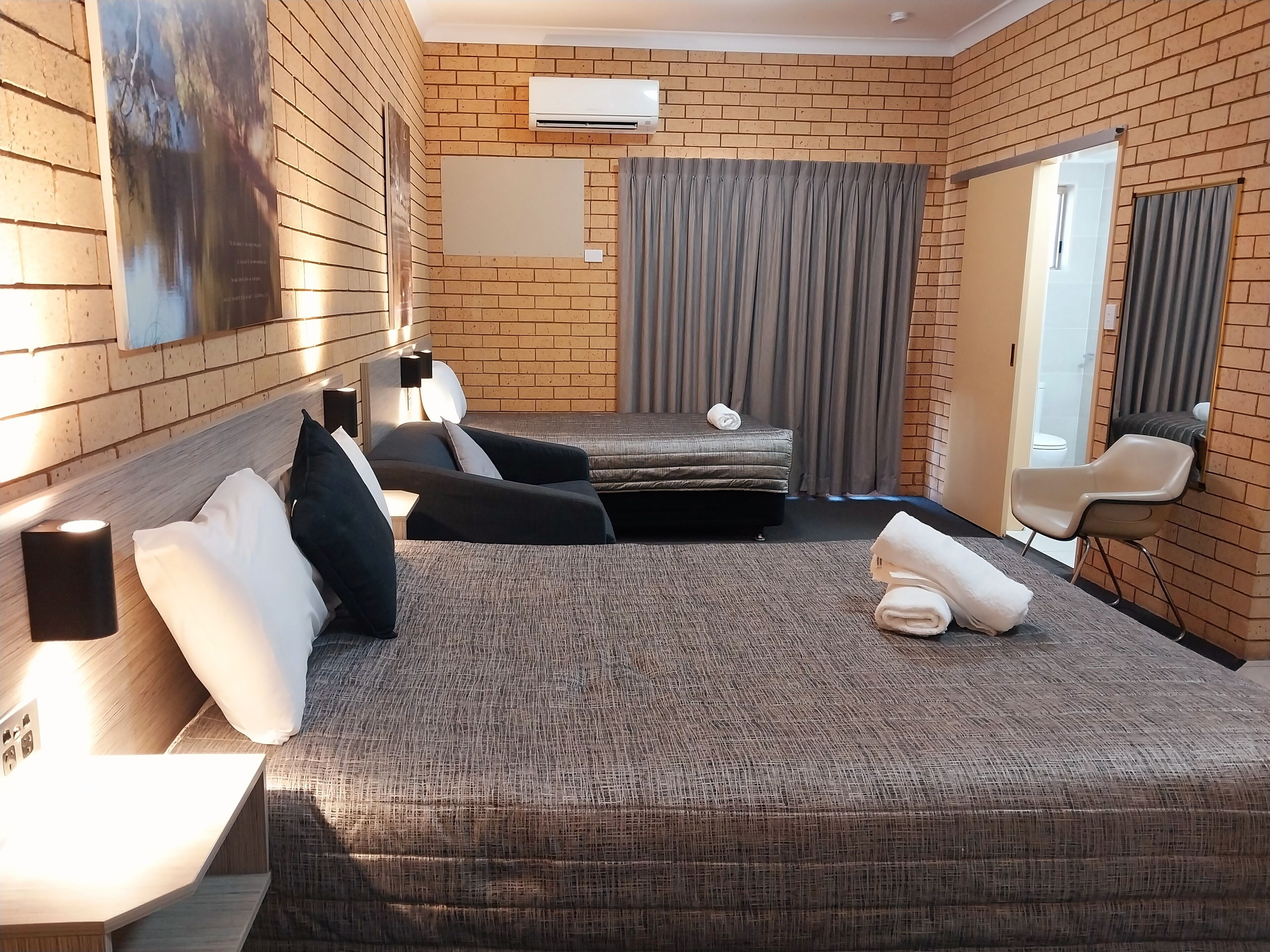Une chambre d’hôtel avec un mur en briques, un lit doté d’une tête de lit rembourrée de couleur grise, un petit canapé, un bureau avec une lampe et une fenêtre avec des rideaux.