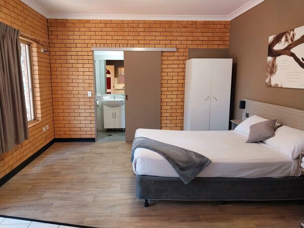 Un dormitorio con pared de ladrillo, una cama con almohadas, un armario y un baño visible a través de una puerta abierta.