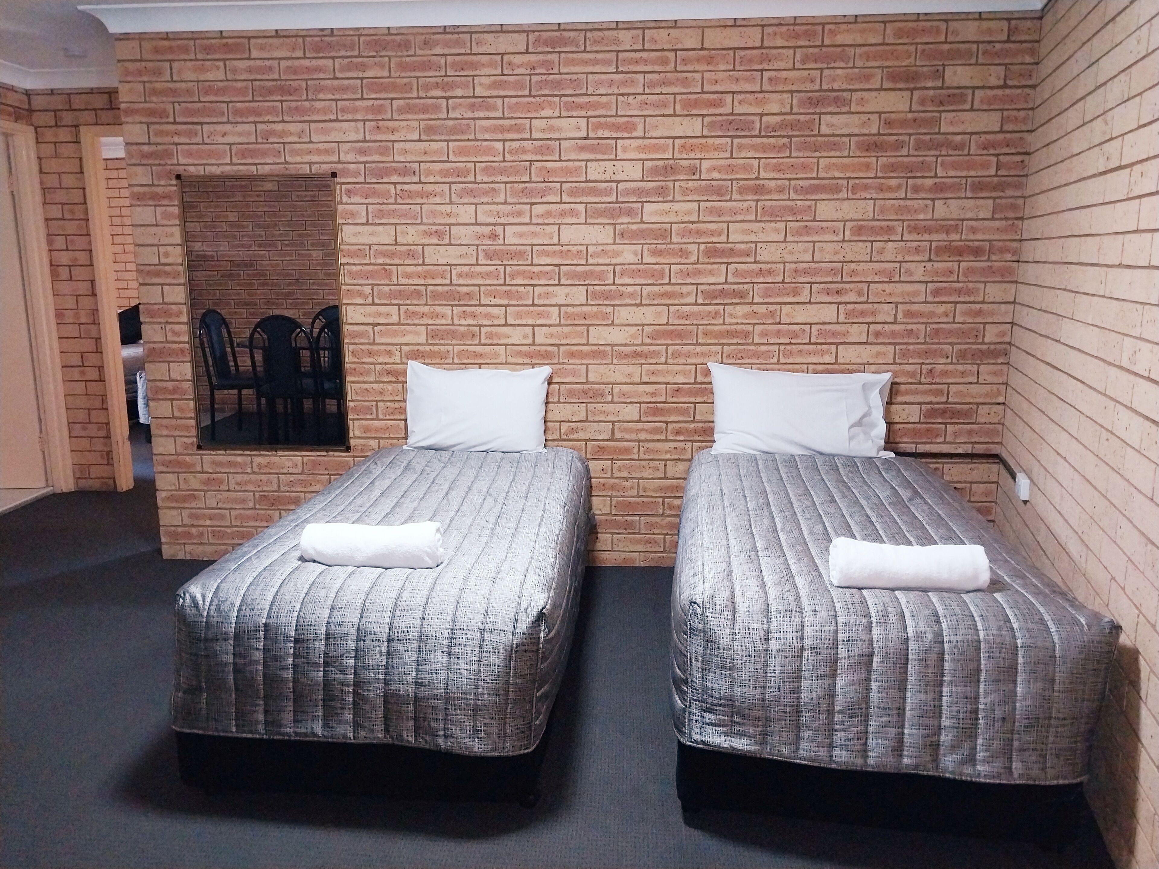 A room with two beds, each with a white pillow and a folded towel. The beds have grey striped bedding. The room has a brick wall and a window with a view of chairs outside.