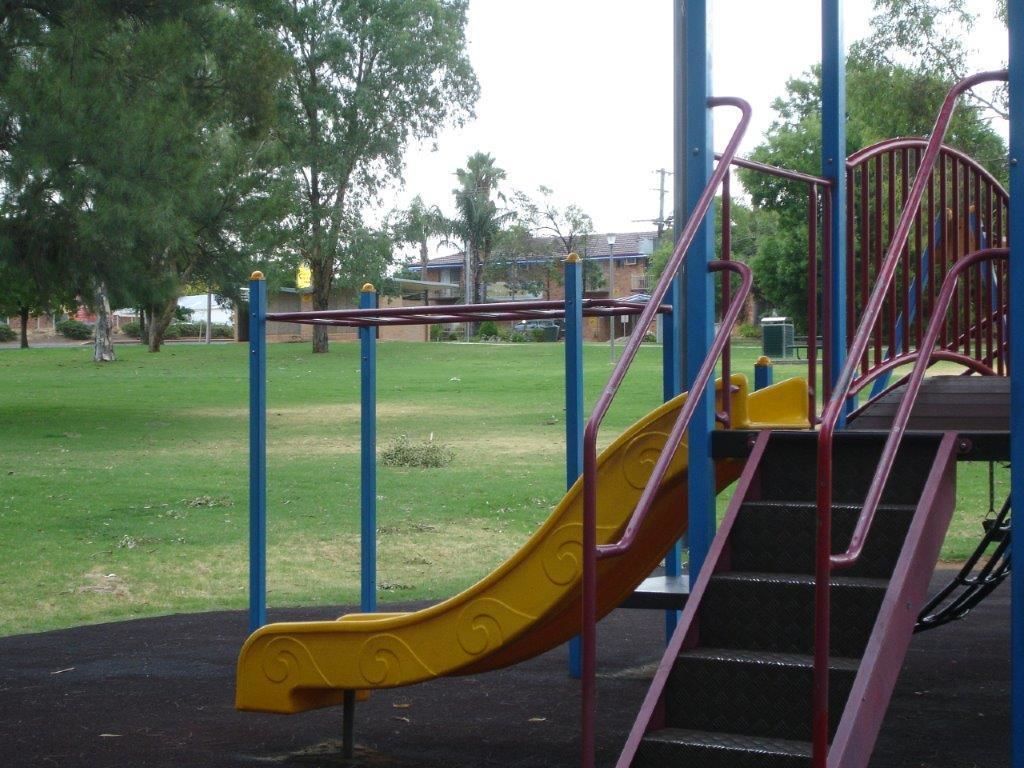 Un parque infantil con un tobogán amarillo, estructuras de escalada azules y rosas, y una zona verde con árboles.