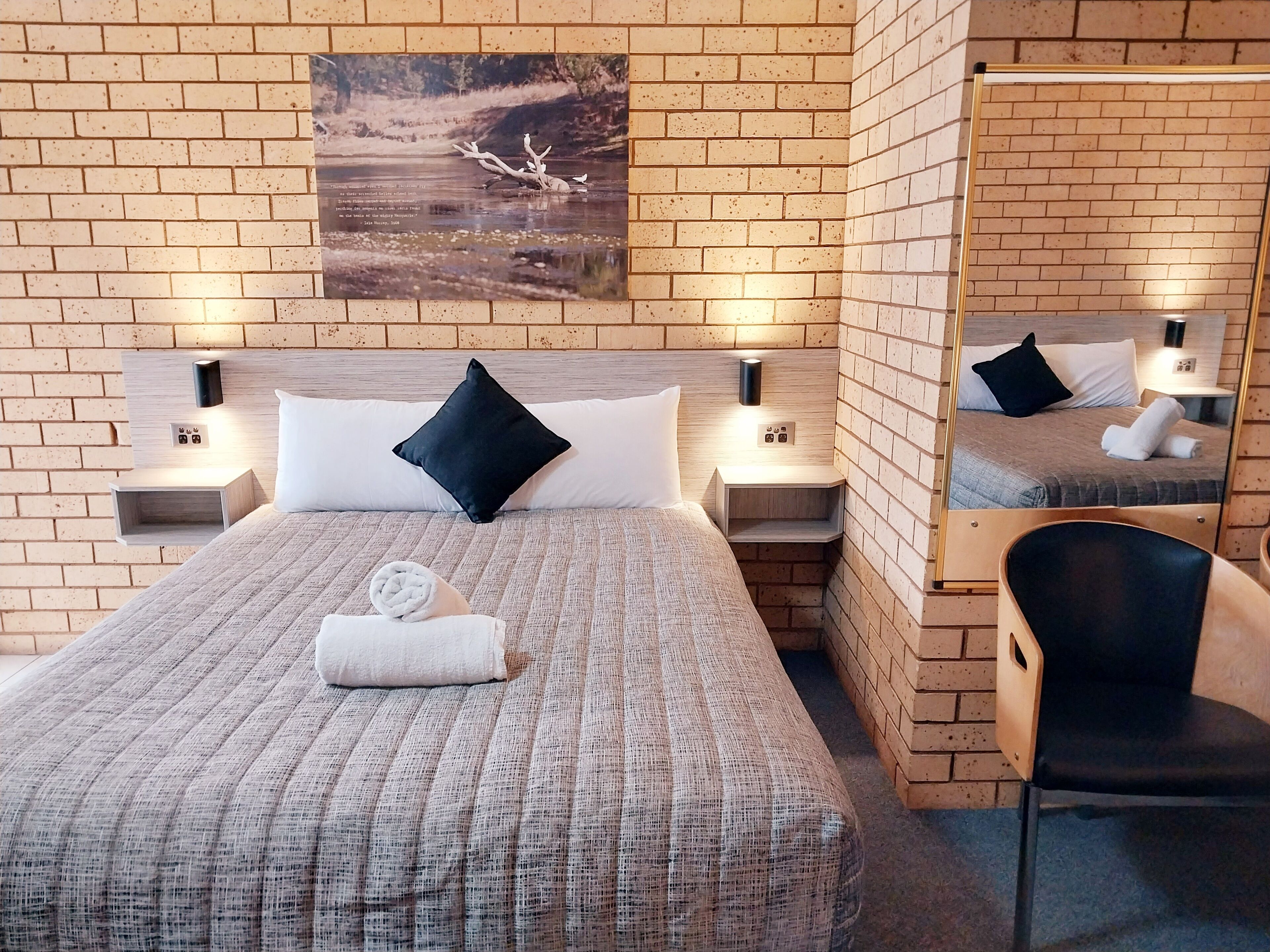 A hotel room with a brick wall, a bed with a textured bedspread, two black pillows, a white towel, and a painting above the bed.