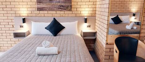 A hotel room with a brick wall, a bed with a textured bedspread, two black pillows, a white towel, and a painting above the bed.
