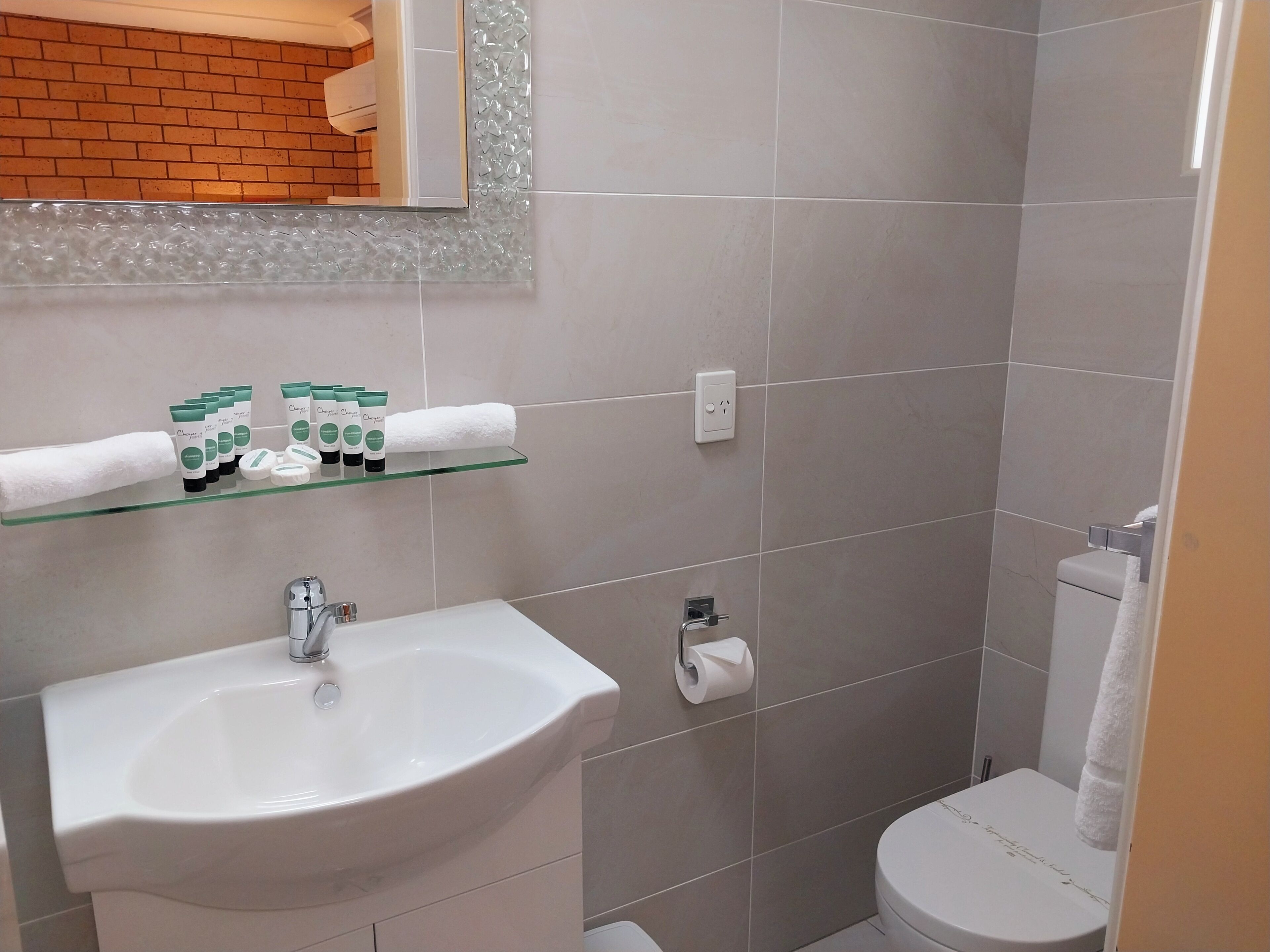 A bathroom with a white sink, a glass shelf with toiletries, a toilet, and a mirror with a brick wall behind it.