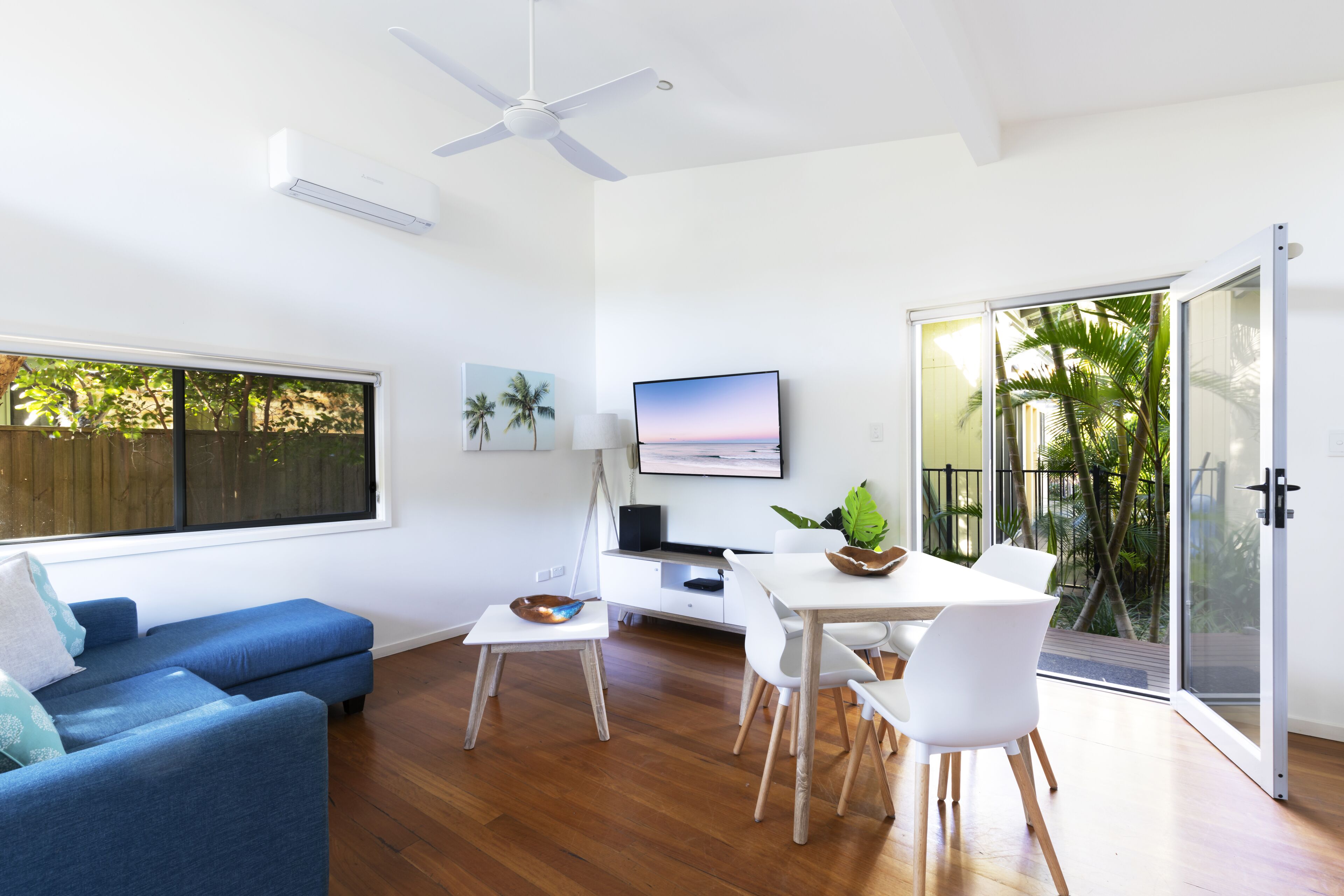 A modern living room with a blue sofa, a dining table with chairs, a flat-screen TV, and a sliding glass door leading to a balcony with plants.