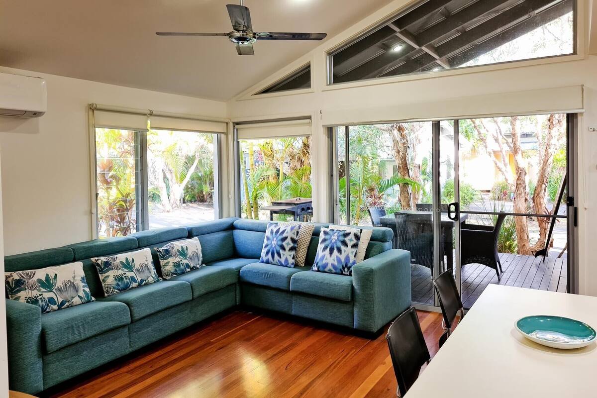 A modern living room with a sectional sofa, a ceiling fan, and large windows offering a view of the outdoors.