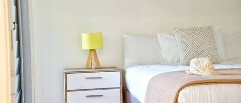A bedroom with a bed, a bedside table with a lamp, a small drawer, and a hat on the bed.