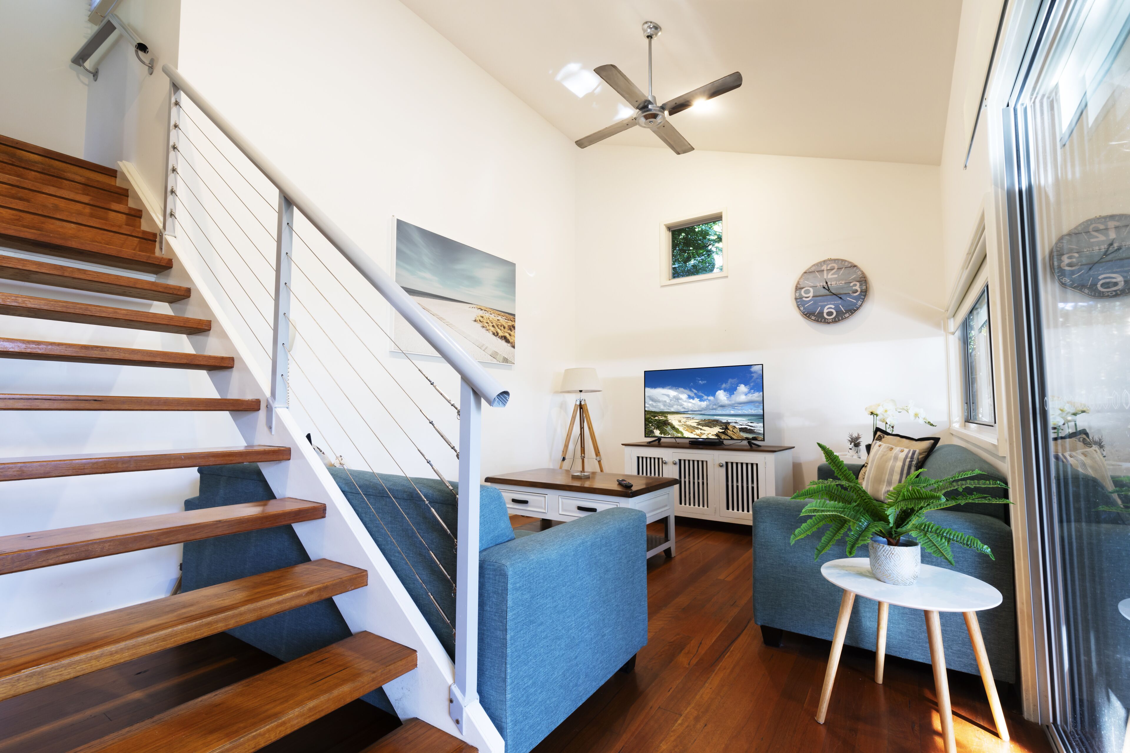 A modern living room with a wooden staircase, a ceiling fan, a sofa, a coffee table, and a potted plant.