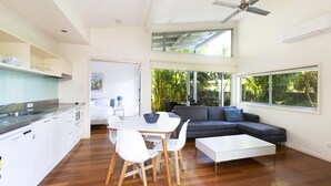 A modern kitchen with white cabinets, a dining area with a white table and chairs, a gray sofa, and large windows offering views of greenery. - Mobys Beachside Retreat (Boomerang Beach)