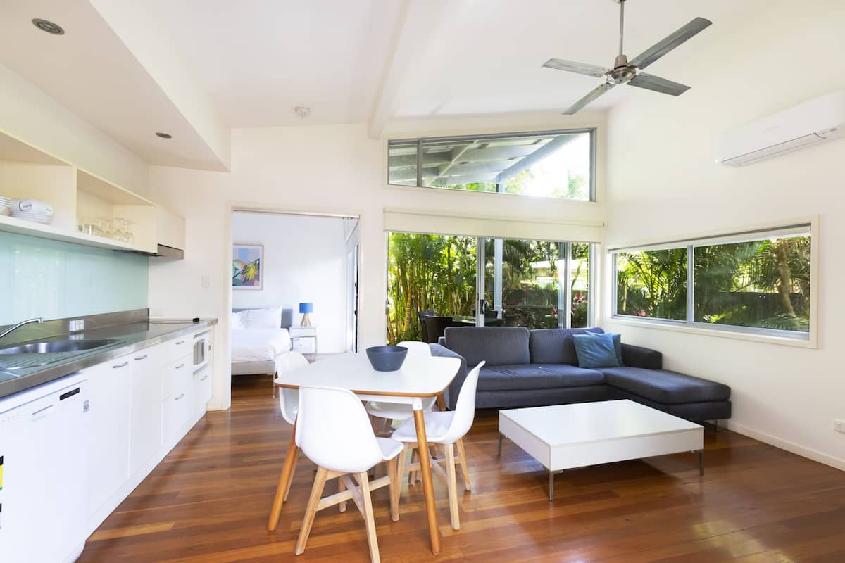 A modern kitchen with white cabinets, a dining area with a white table and chairs, a gray sofa, and large windows offering views of greenery.
