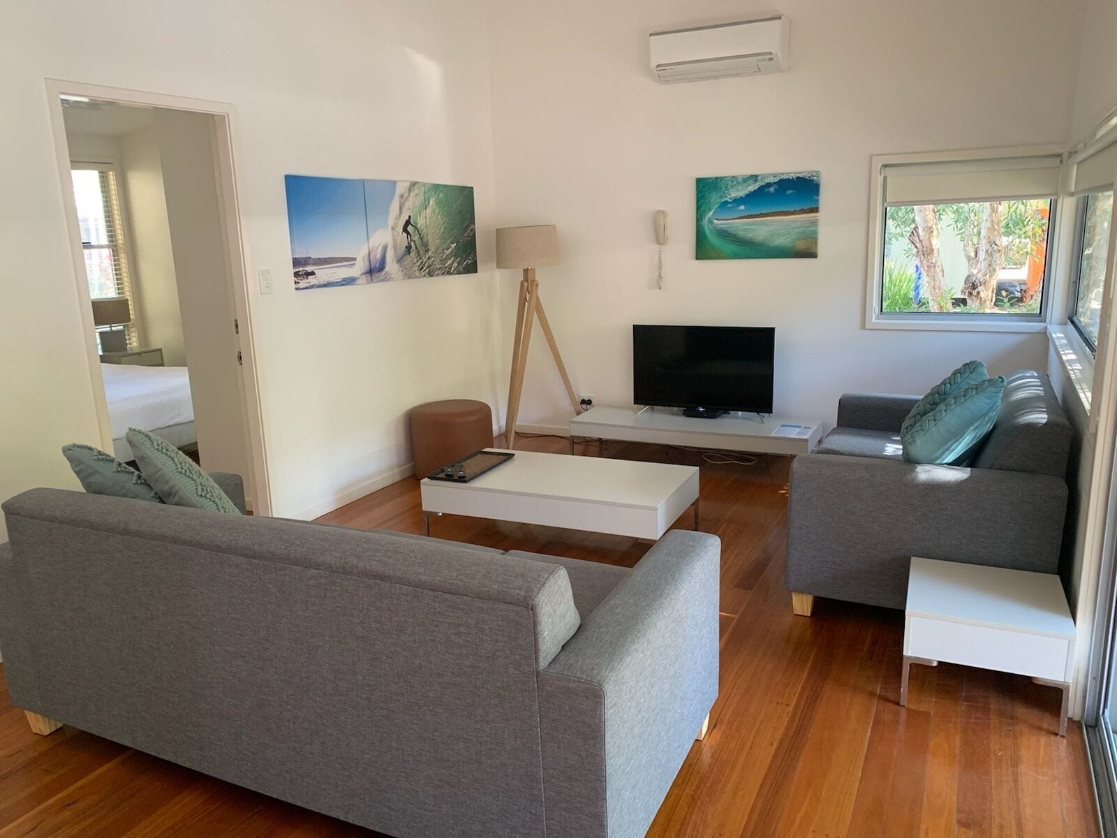 A modern living room with a grey sofa, a smaller grey armchair, a white coffee table, and a TV on a white stand. There are two paintings on the wall, a lamp, and a window with a view of greenery.