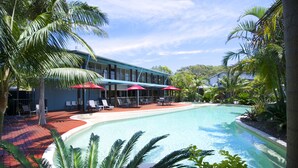 A pool area with lounge chairs and umbrellas, surrounded by palm trees and a building with large windows. - Mobys Beachside Retreat (Boomerang Beach)